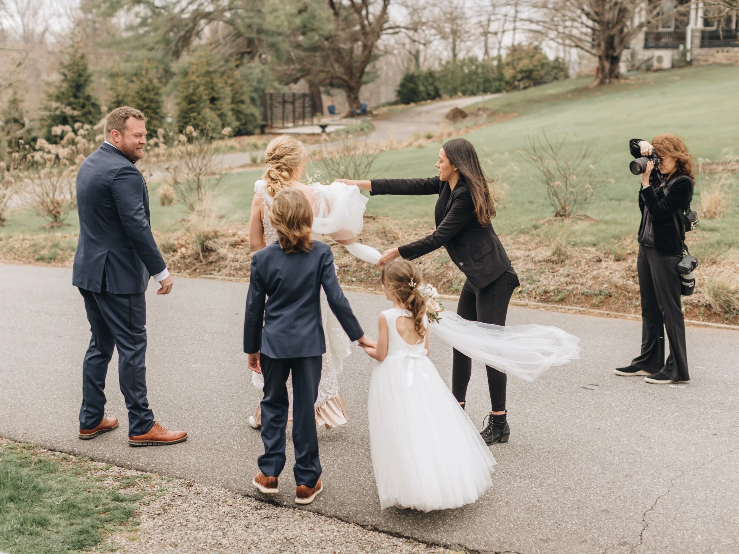 A group of people, including a bride in a wedding dress, a groom, and children, are standing outdoors during a wedding photo shoot. A woman with a camera is photographing the scene.