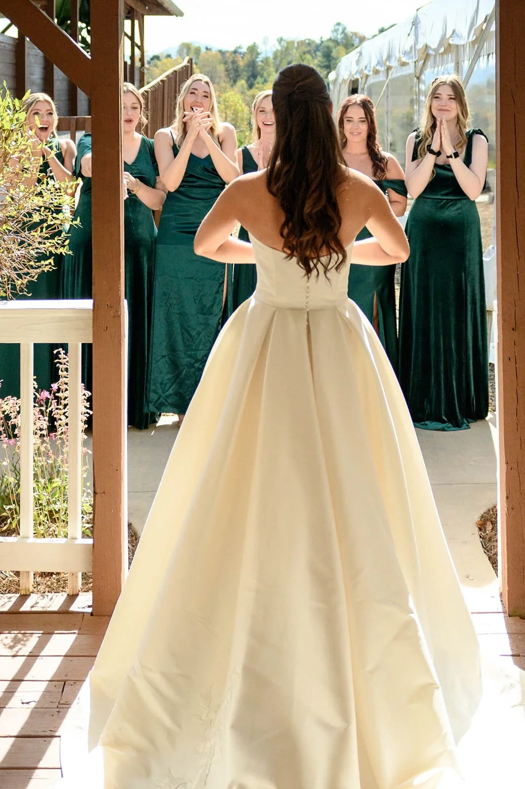 A bride in a cream wedding gown faces her bridesmaids, who are dressed in matching green dresses, outside during a sunny day.