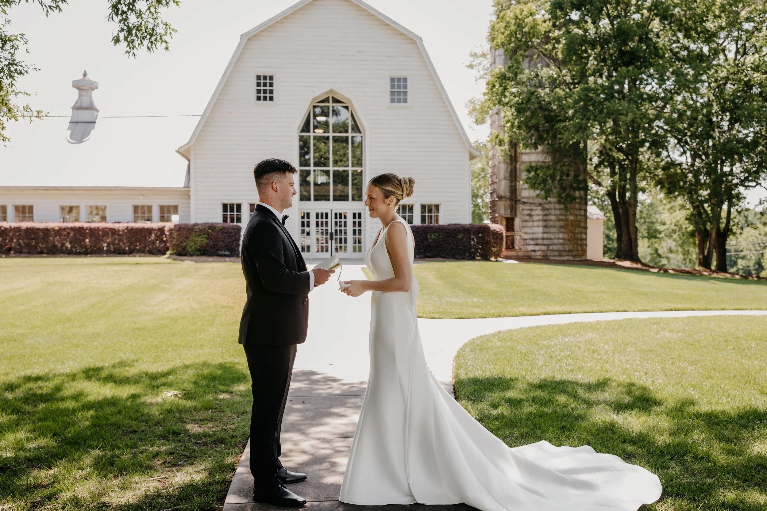 A bride and groom standing on a sidewalk in front of a white chapel, holding hands and facing each other during their wedding ceremony outdoors on a sunny day. The bride is wearing a white gown and the groom is in a black tuxedo.