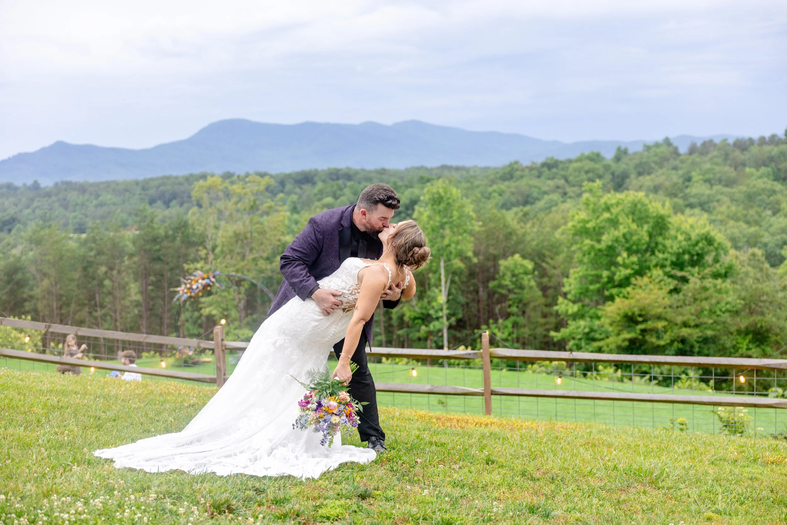 Bride and groom sharing a kiss in a green outdoor setting with trees and mountains in the background, the bride holding a bouquet of flowers.