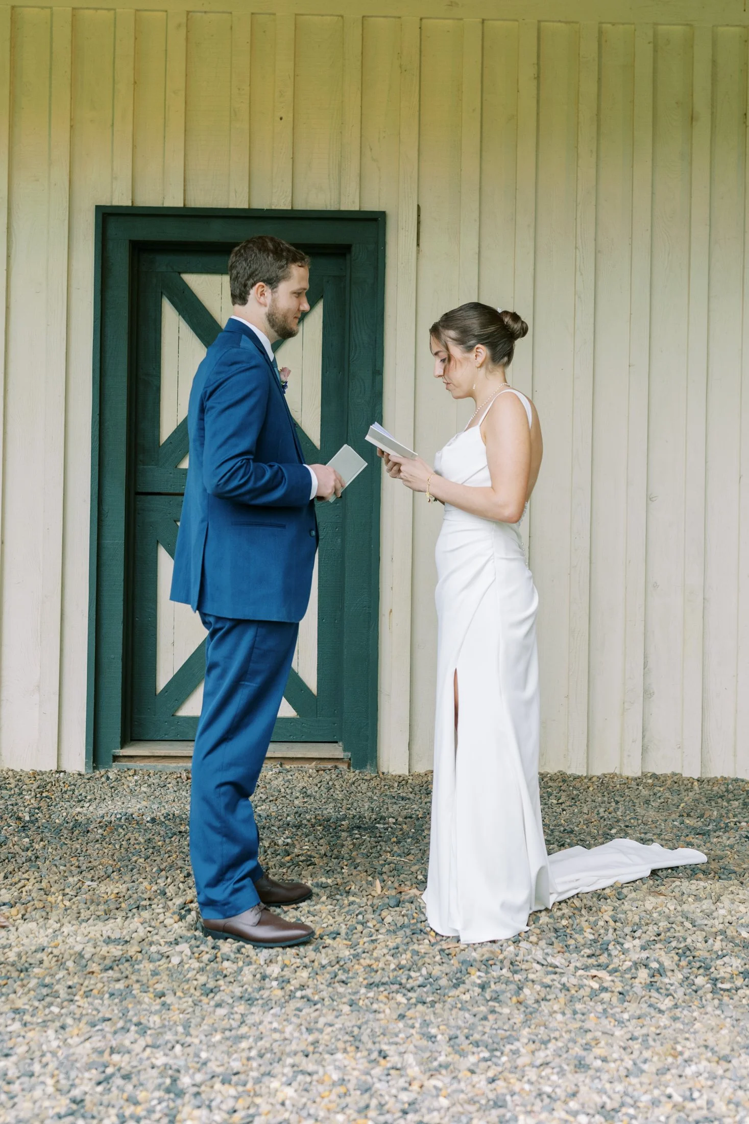 A bride and groom exchange wedding vows outdoors, standing on a gravel surface in front of a wooden wall and door, with both holding booklets.