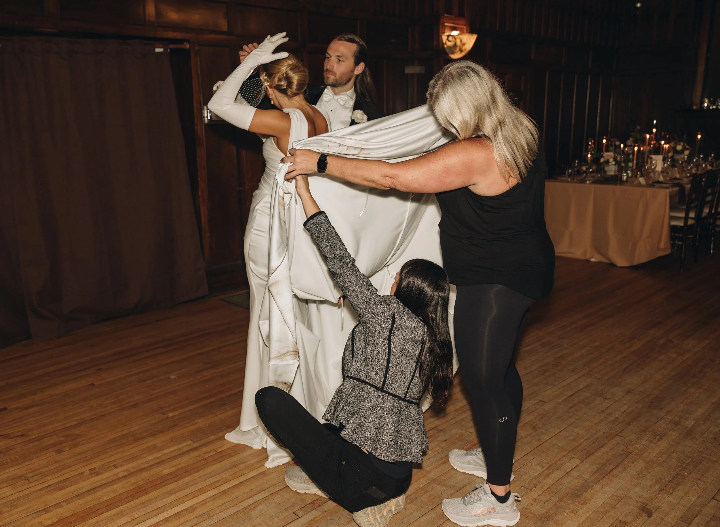 Three women and a man in a wedding dress preparing for a First Look moment at a wedding reception, with one woman adjusting her dress, the bride getting ready, and the other woman assisting, in a wooden-floored event hall.