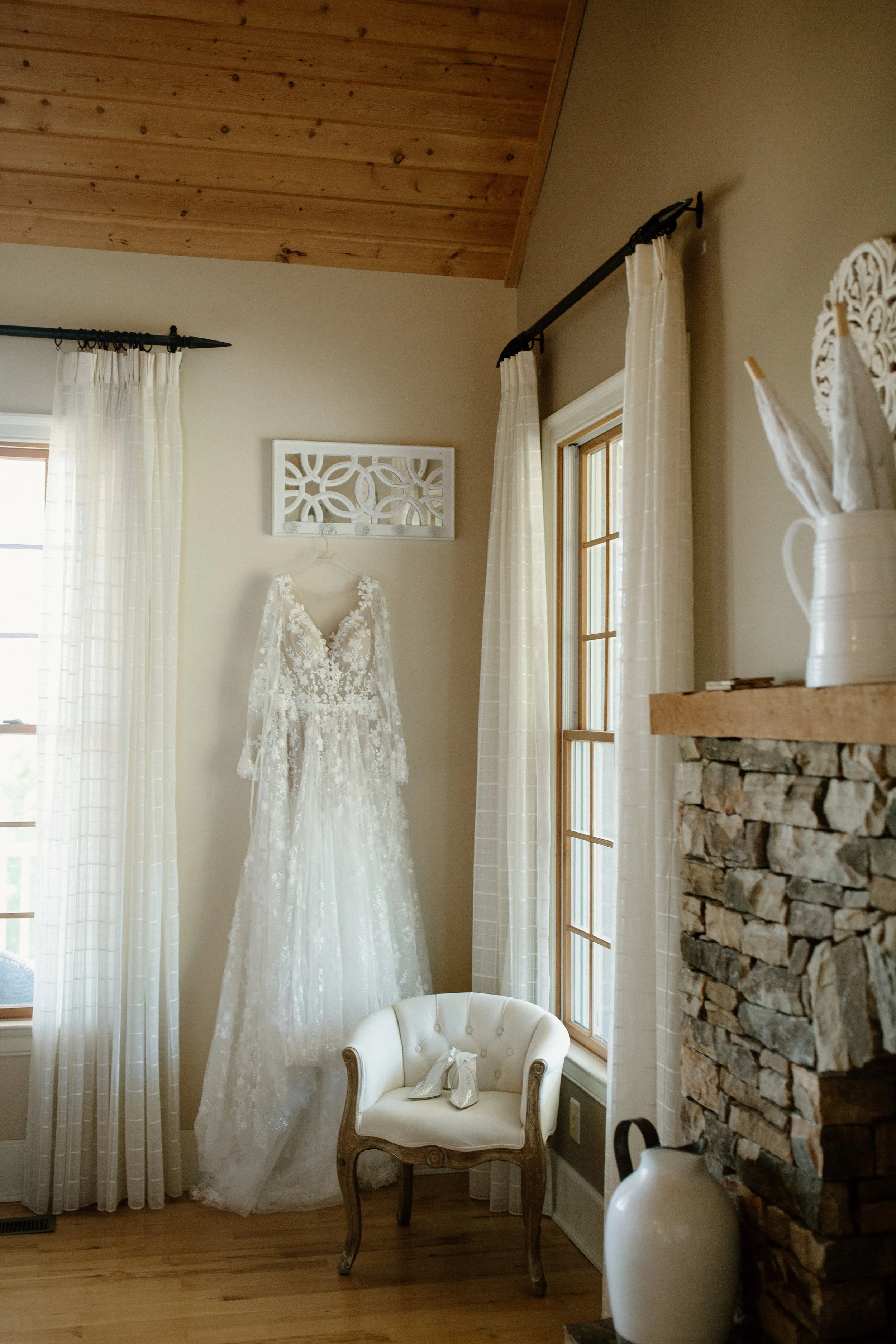 A wedding dress hanging on a wall next to a window and a small vintage armchair with lace shoes on it, in a cozy room with a stone fireplace and wooden ceiling.