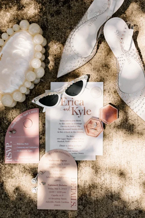 Wedding invitation, pearl jewelry, white sunglasses with rhinestones, a pair of fancy shoes, and RSVP and informational cards for Erica and Kyle's wedding.