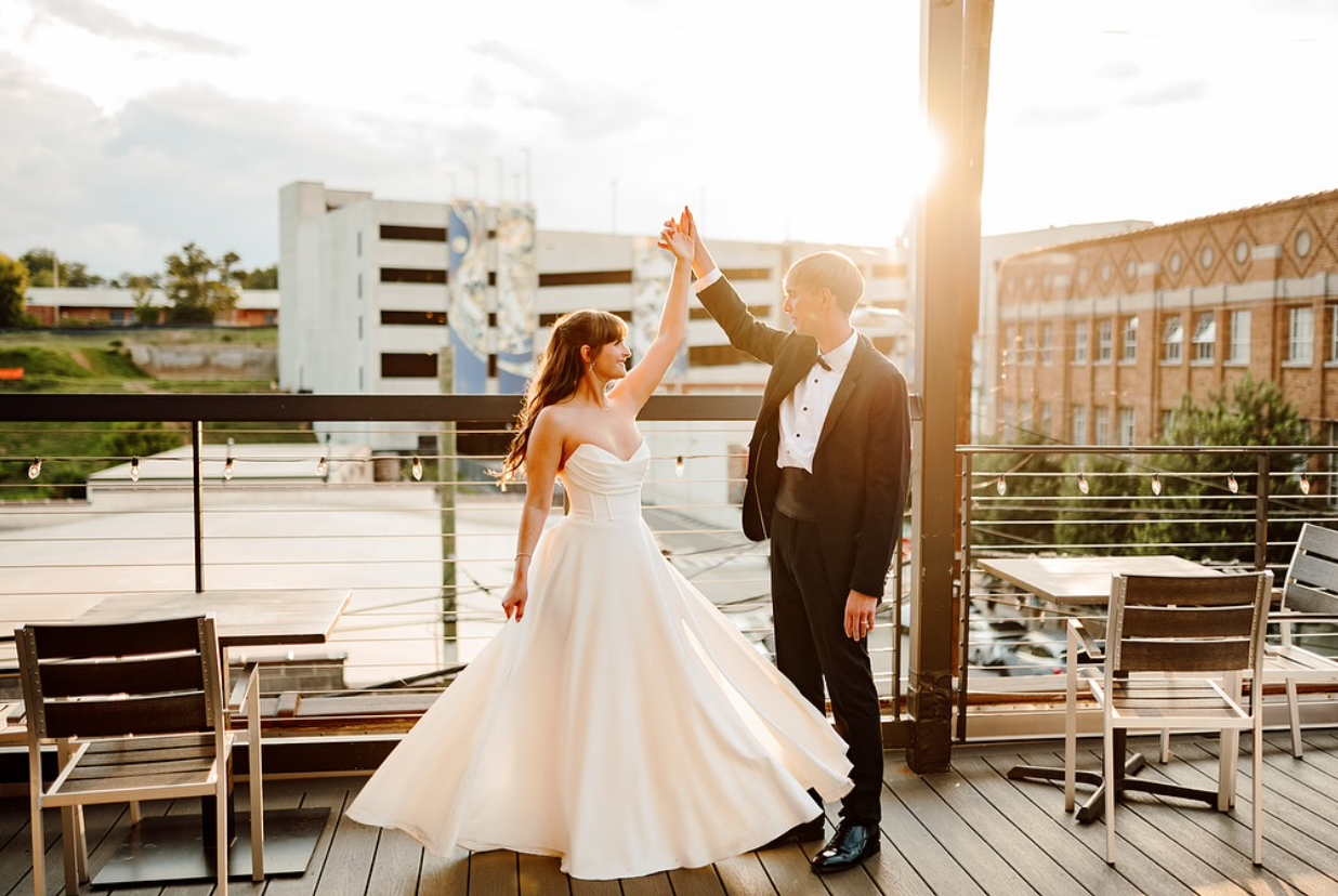 Bride and groom dancing on a balcony during sunset, holding hands, with outdoor furniture and city buildings in the background.