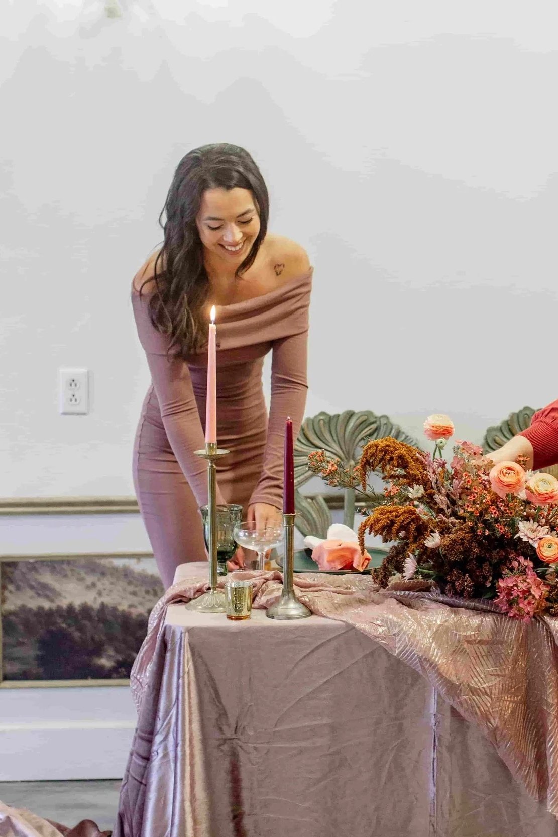 A woman with long dark hair smiling and leaning over a table decorated with pink and orange flowers, candles, and glasses, during a celebration or gathering.
