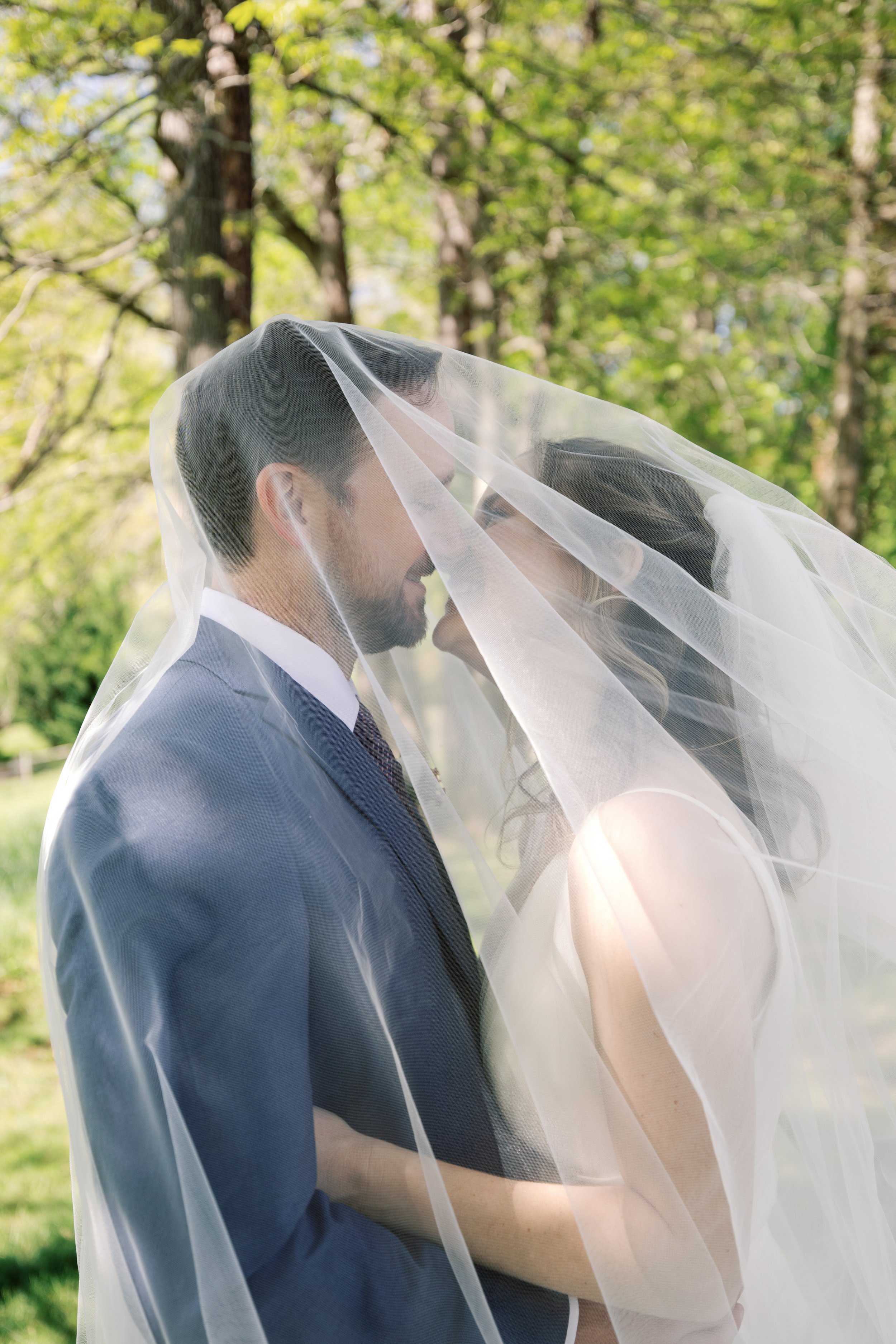 A bride and groom under a veil in a park with green trees in the background.