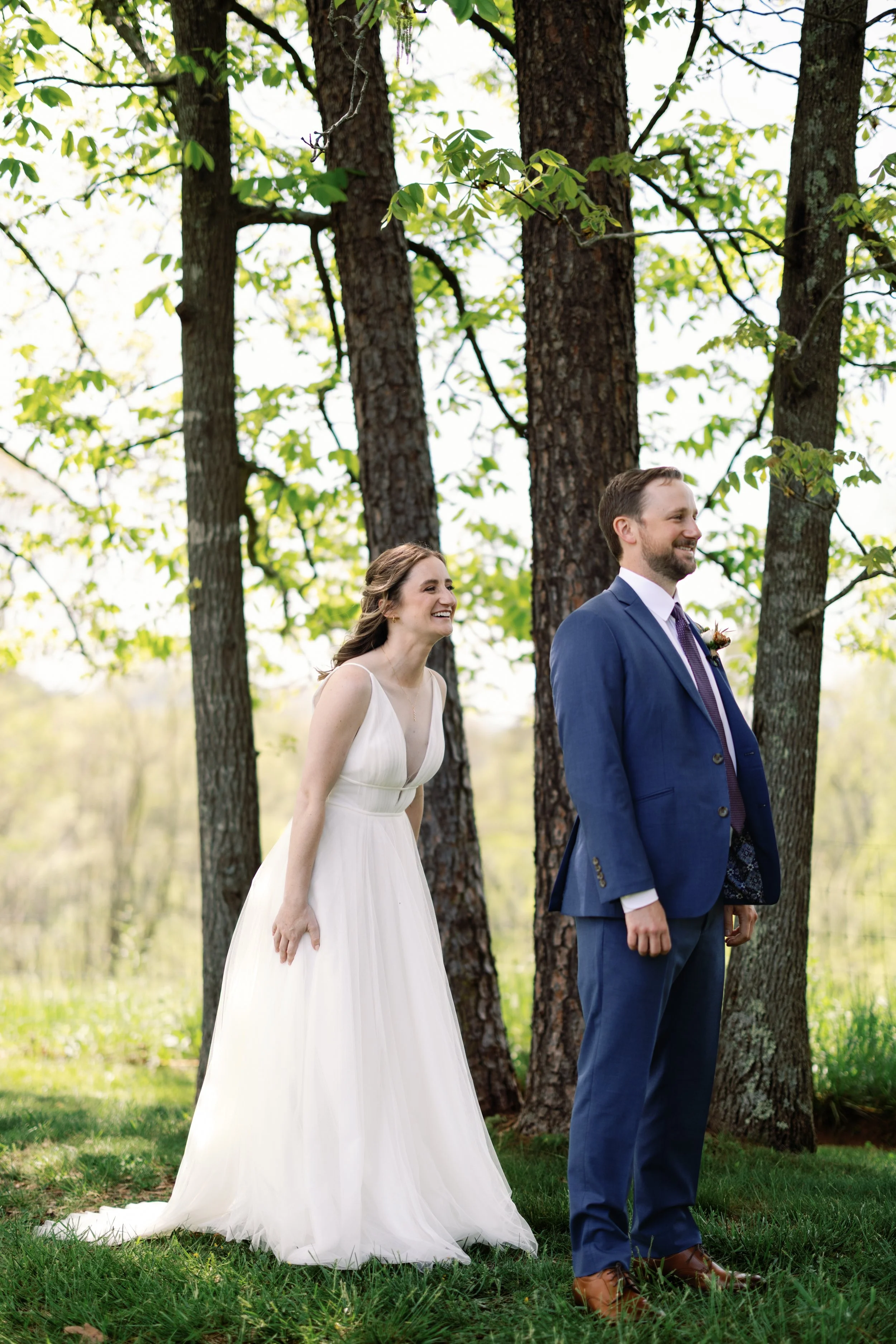 Bride in a white wedding dress and groom in a blue suit standing outdoors near trees, smiling at each other.