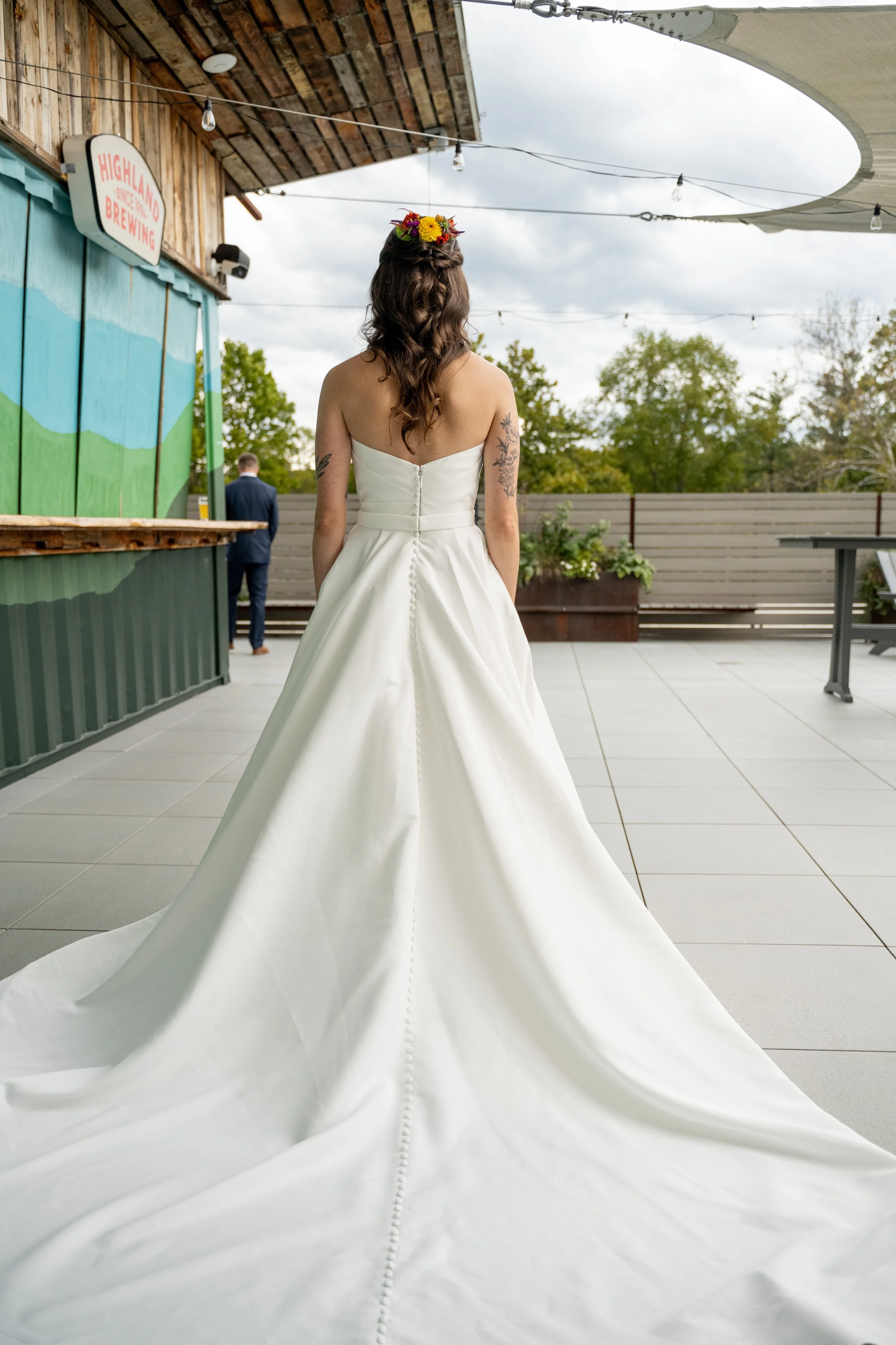 A woman in a white wedding dress with a long train standing on a patio, facing away from the camera, with a flower crown in her hair.