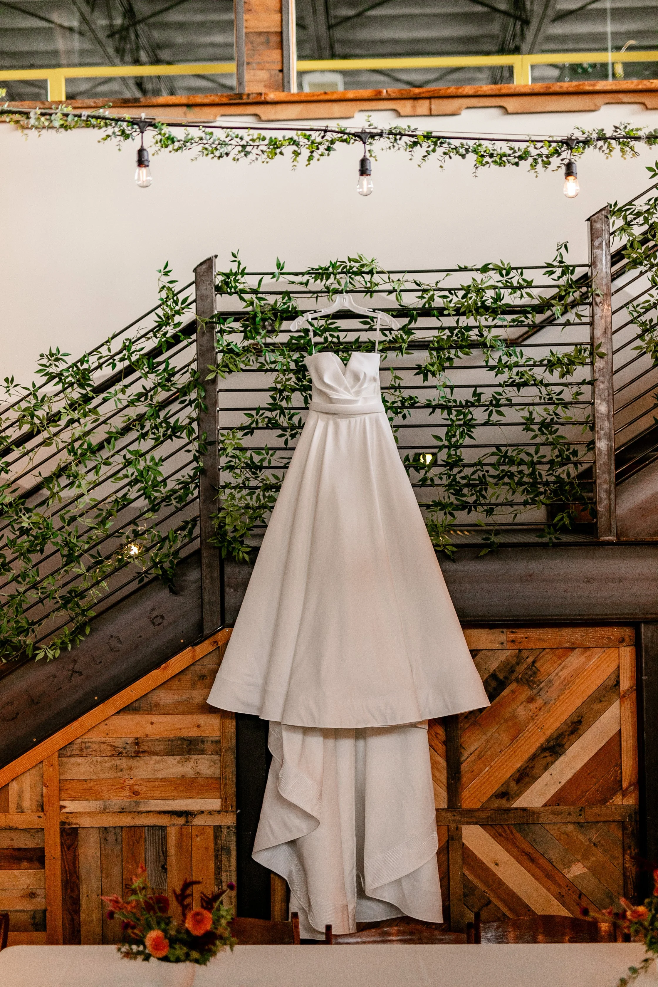 A wedding dress hanging on a wall with greenery decorations on a metal railing, and string lights overhead.