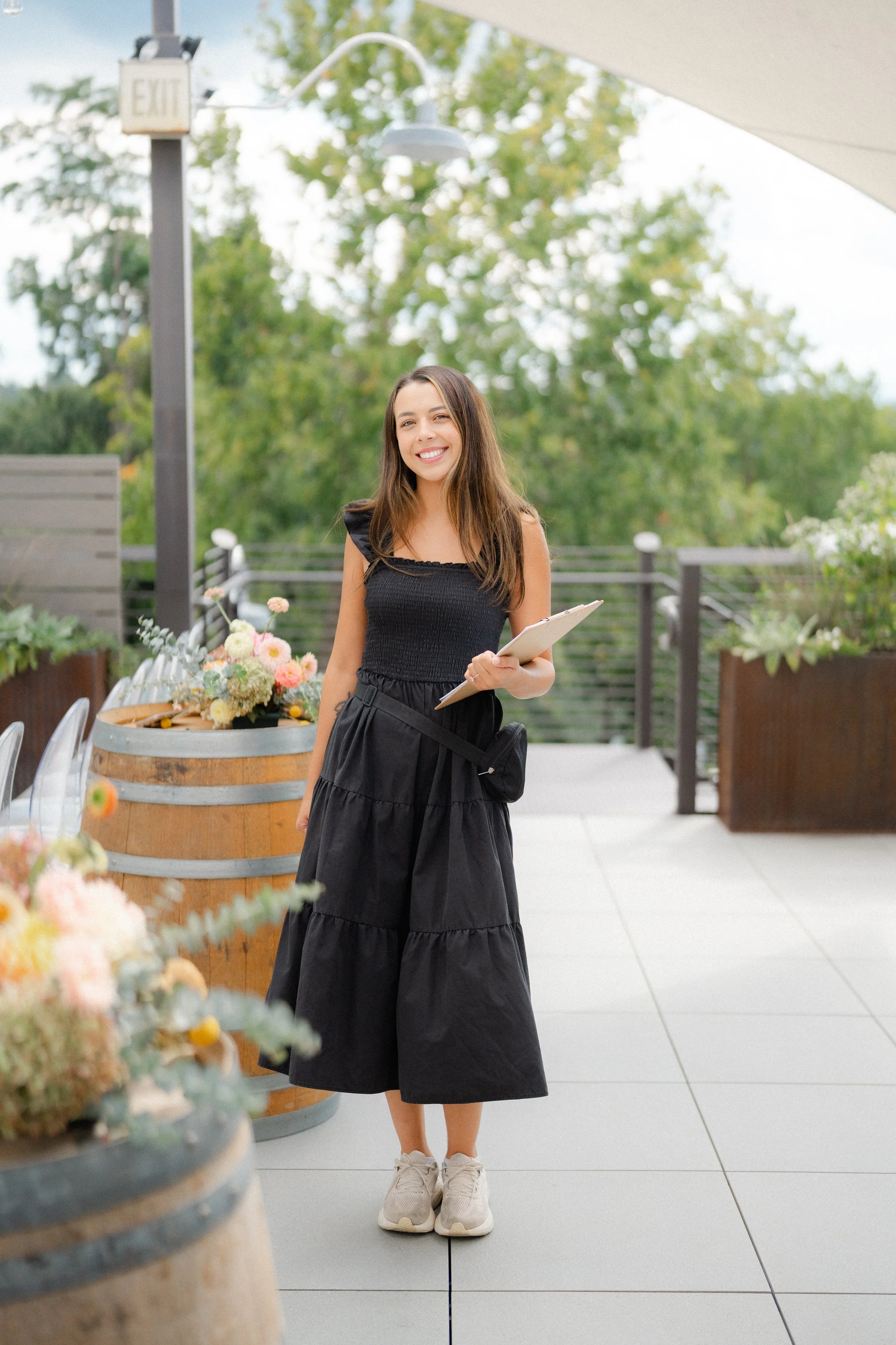 A young woman in a black dress and sneakers stands outside on a patio, smiling and holding a notepad. There are flower arrangements in barrels nearby, and greenery in the background.