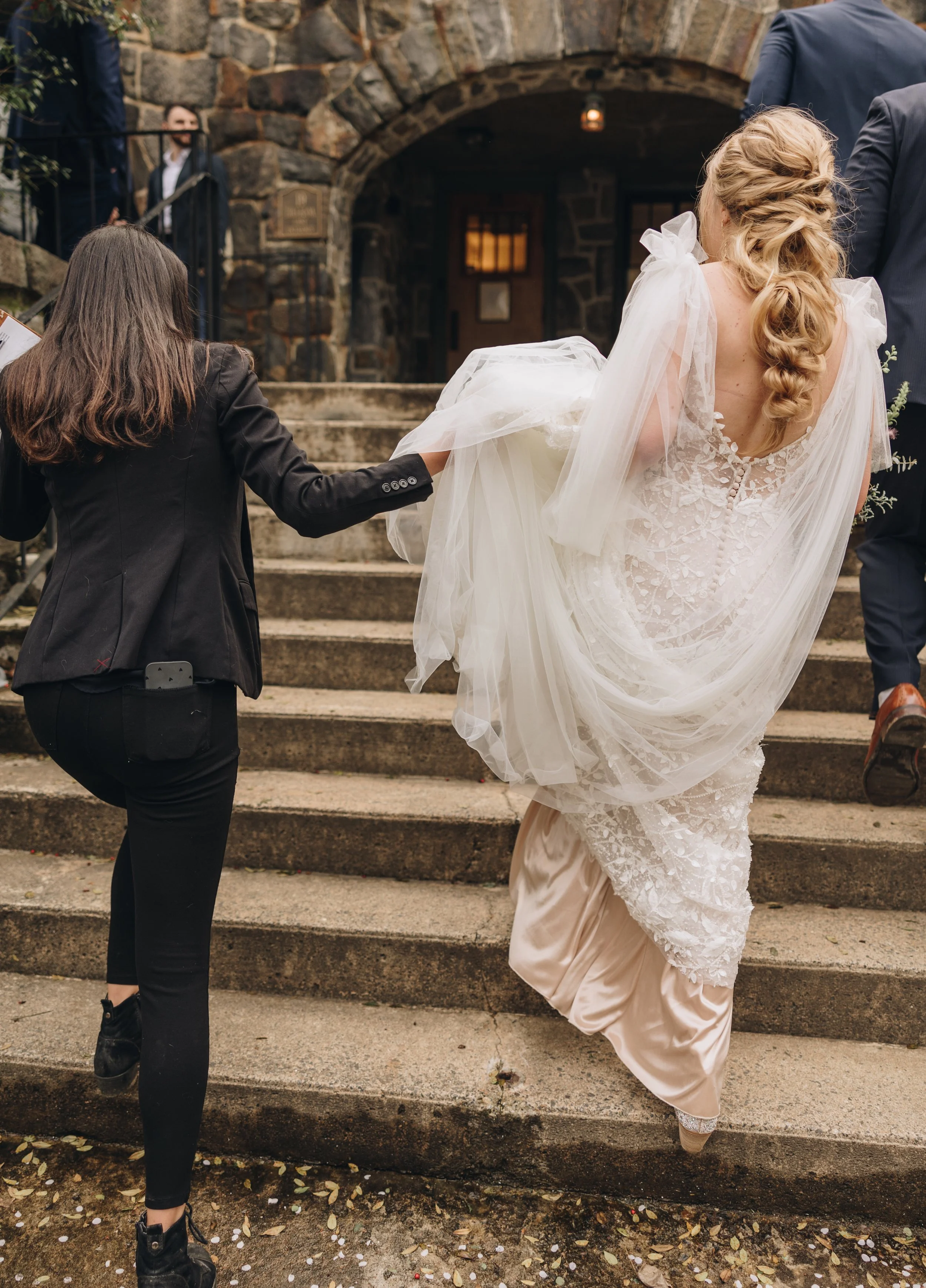 A bride with long, curly blonde hair in a wedding gown is ascending outdoor stone steps, assisted by a woman in a black suit. The scene is outside a stone building with an arched doorway, and other guests are visible in the background.