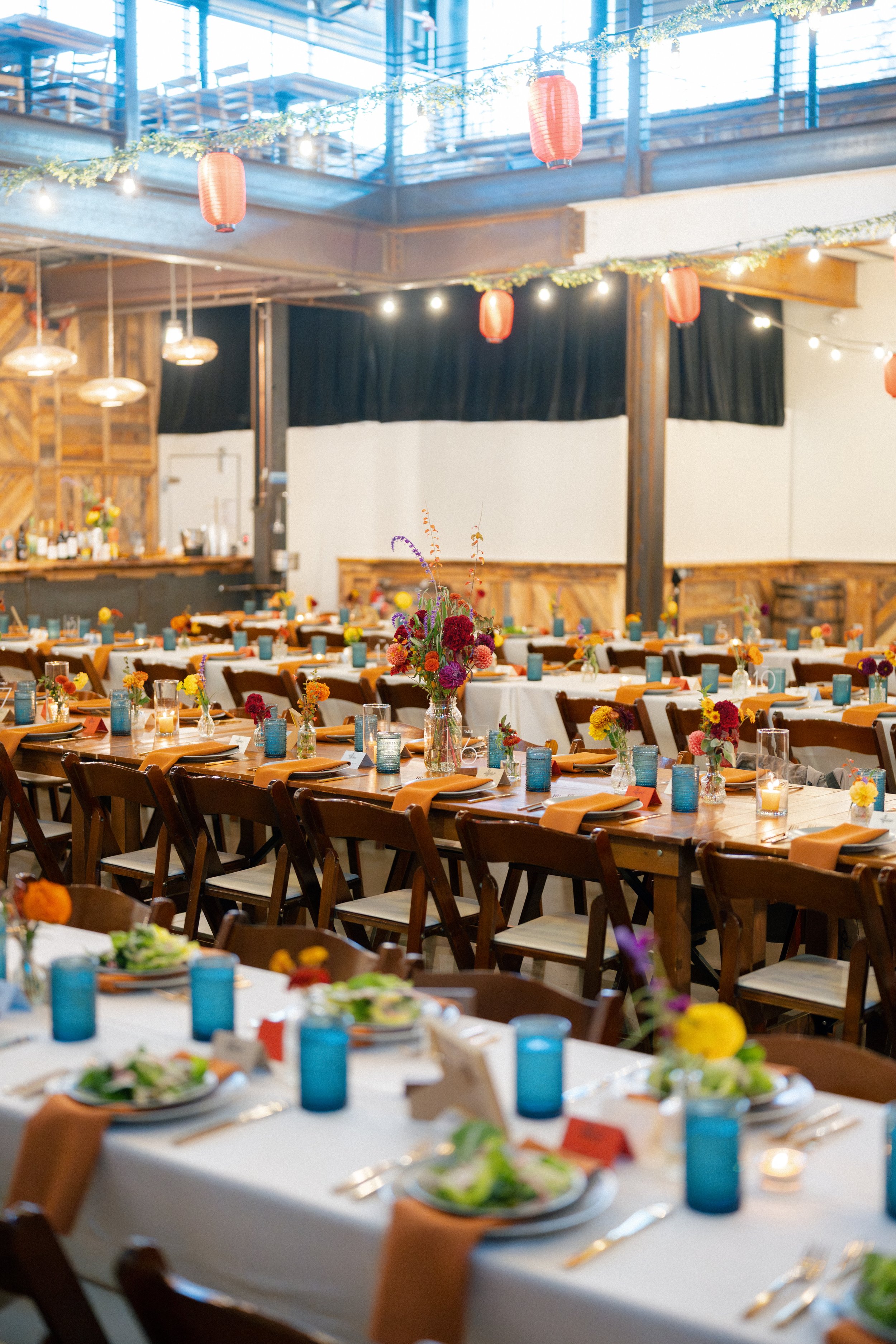 Set dining tables decorated with flowers, candles, and napkins in a rustic venue with string lights and hanging lanterns.