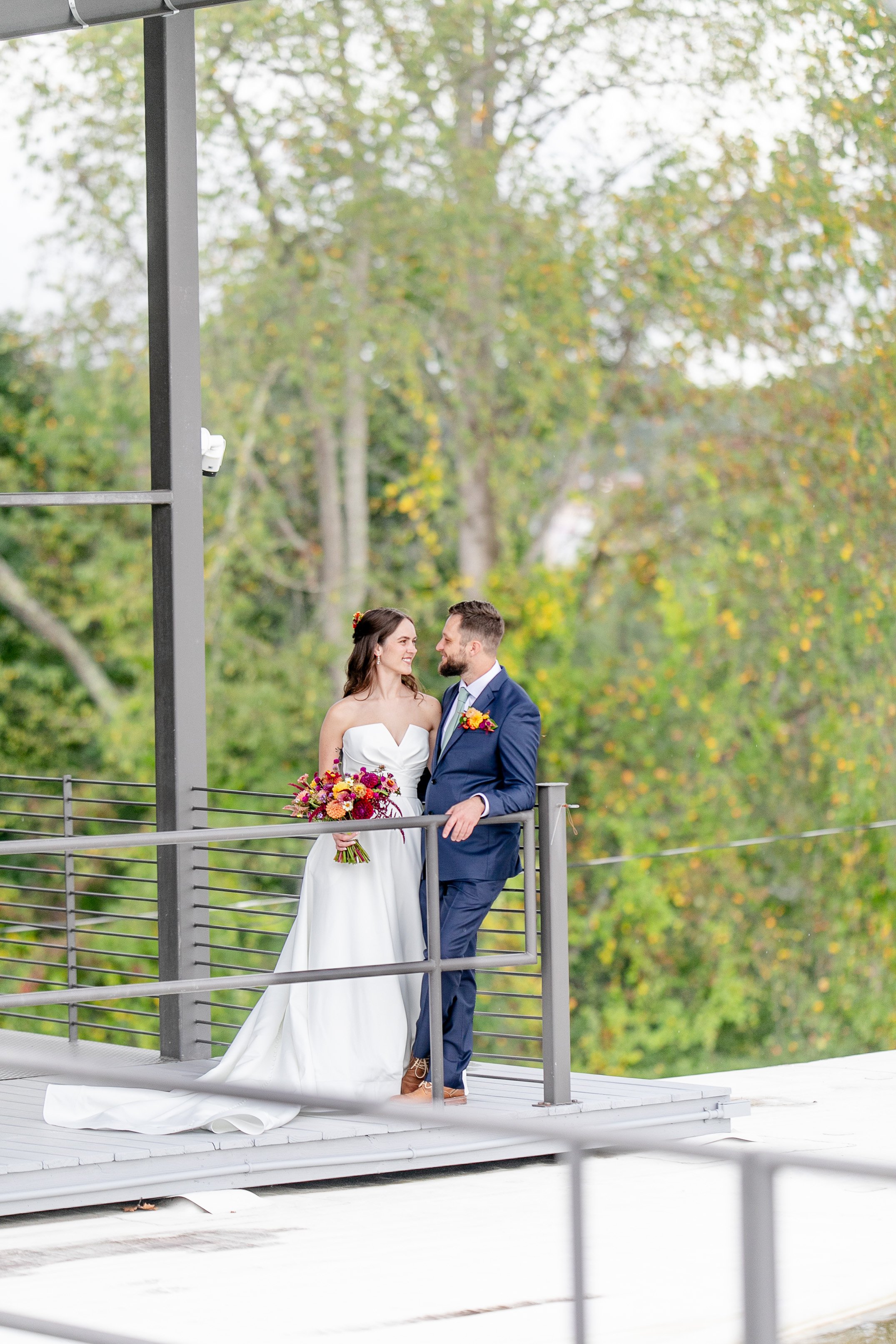 A bride and groom standing on a balcony, gazing at each other, with trees and green foliage in the background during their wedding.