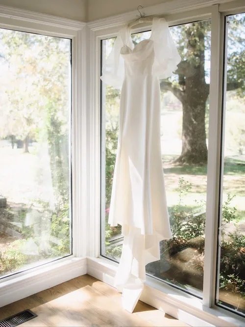 A white wedding dress hanging from the ceiling near large corner windows with a view of trees and a sunny outdoor garden.