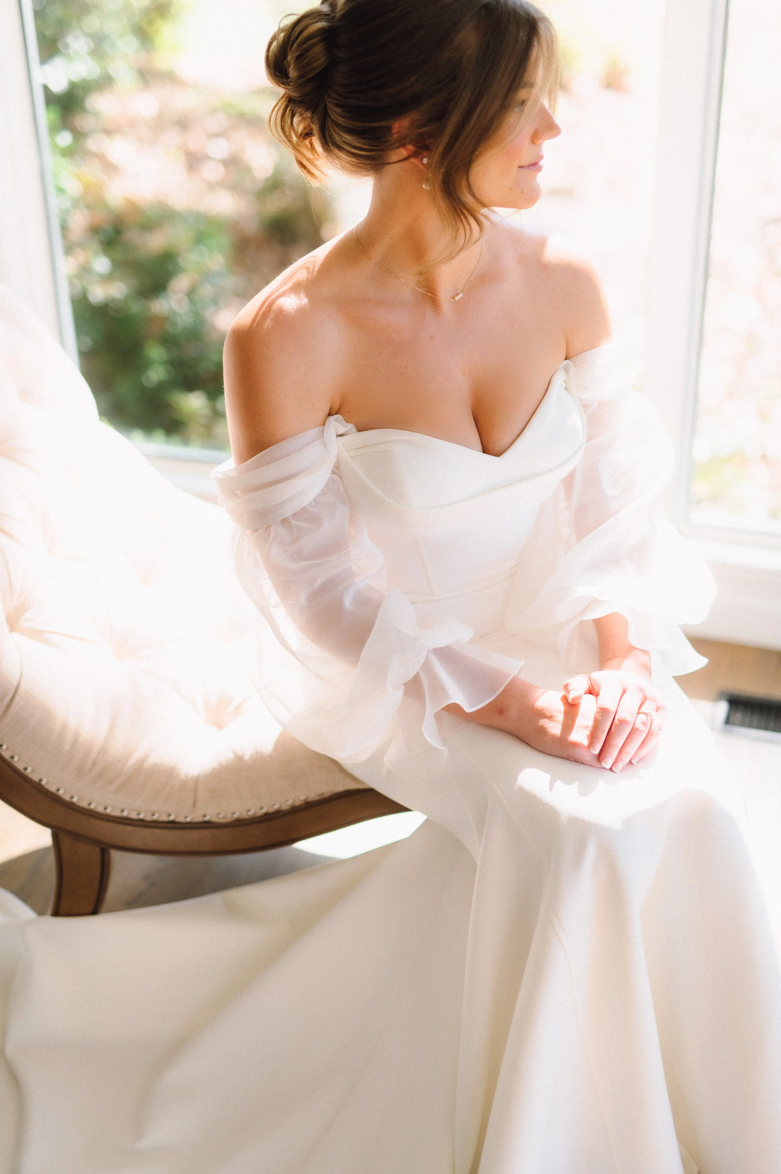 A woman in a white wedding dress sitting on a vintage beige chair by a window, with sunlight illuminating her.