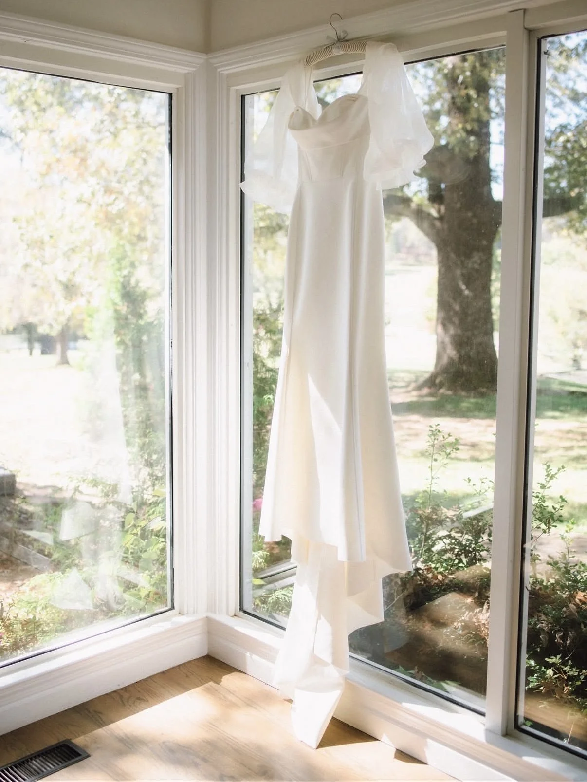 White wedding dress hanging from a hook on the ceiling in front of large window, with trees and greenery outside