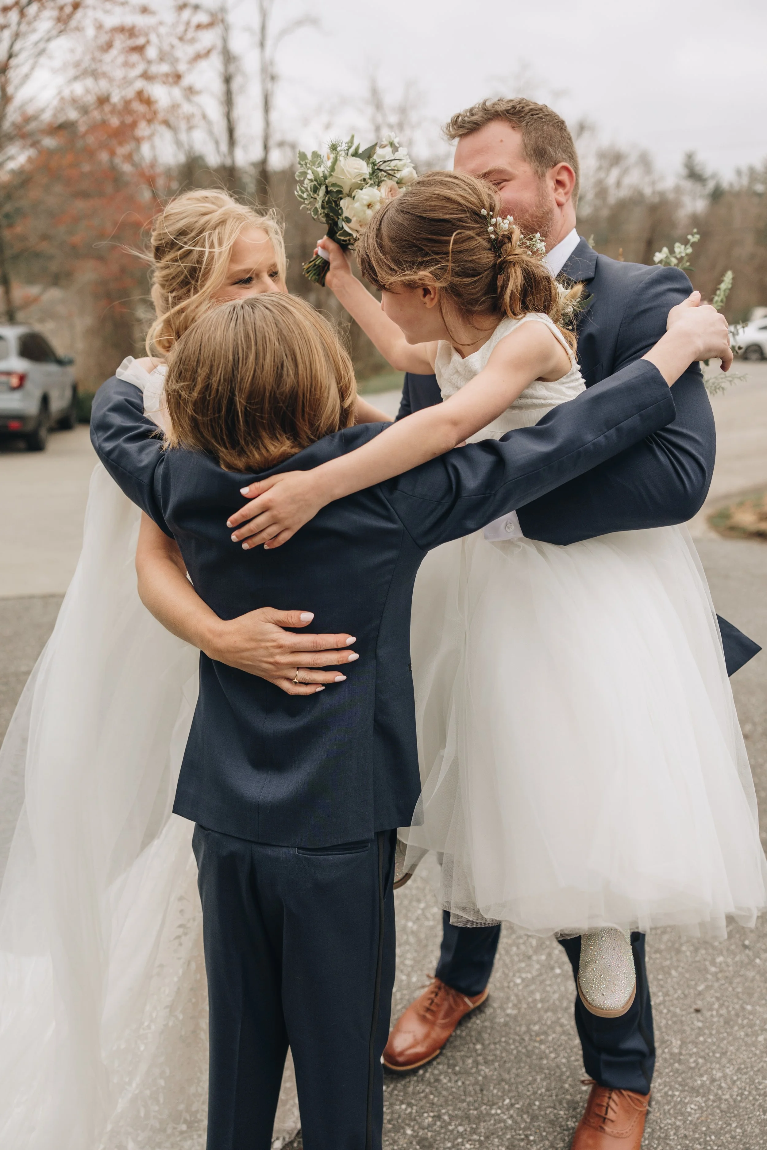 A group of five children, including a bride and groom, embracing each other outdoors in a celebratory hug, with trees and parked cars in the background.