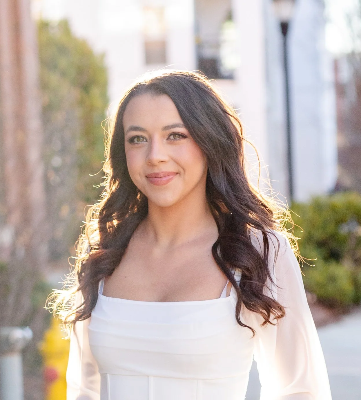A young woman with long, wavy dark hair, wearing a white top, smiling outdoors in sunlight.