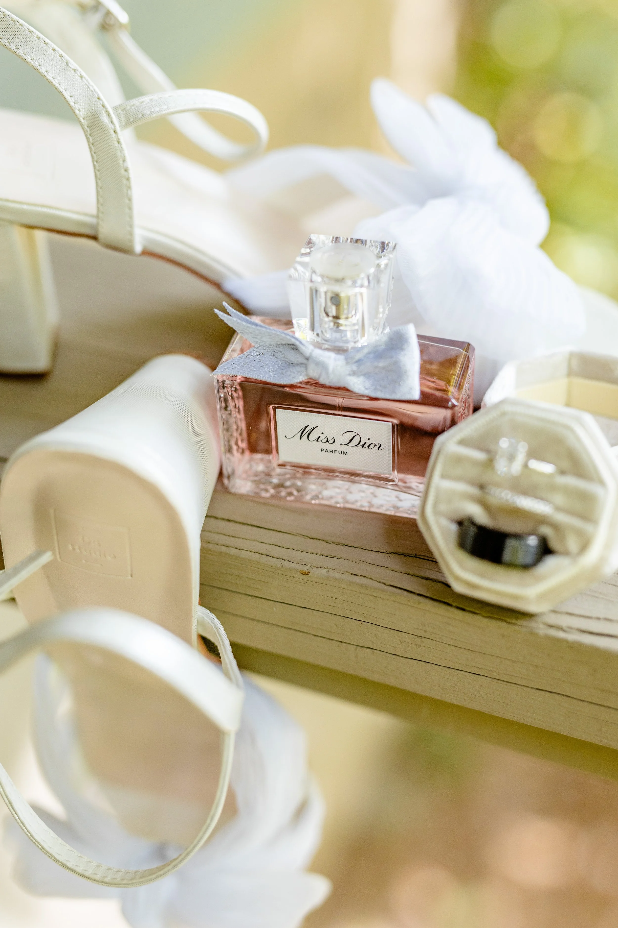 A display of a pink Miss Dior perfume bottle with a silver bow, white shoes, a ring in a velvet ring box, and a white flower on a wooden surface.