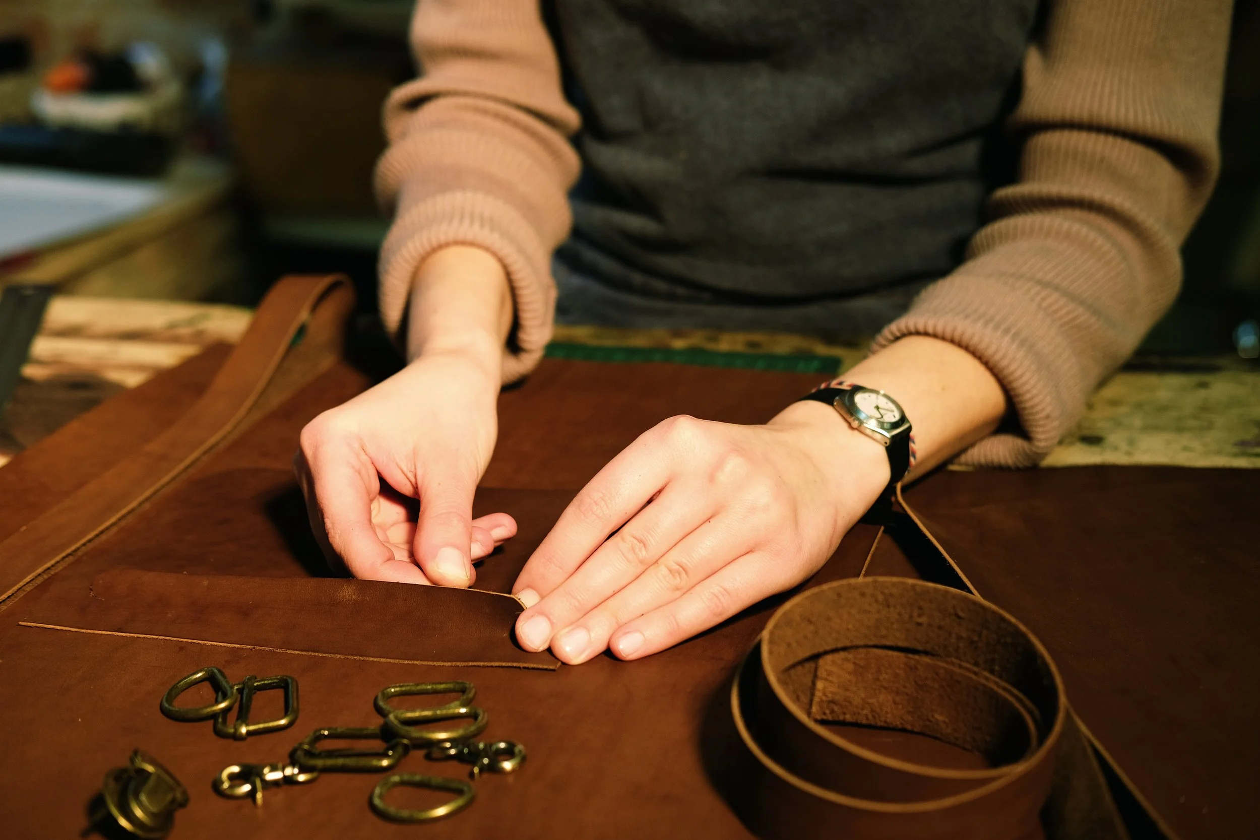 Une personne assemble du cuir marron pour fabriquer une ceinture, avec des boucles en métal posées sur la table.
