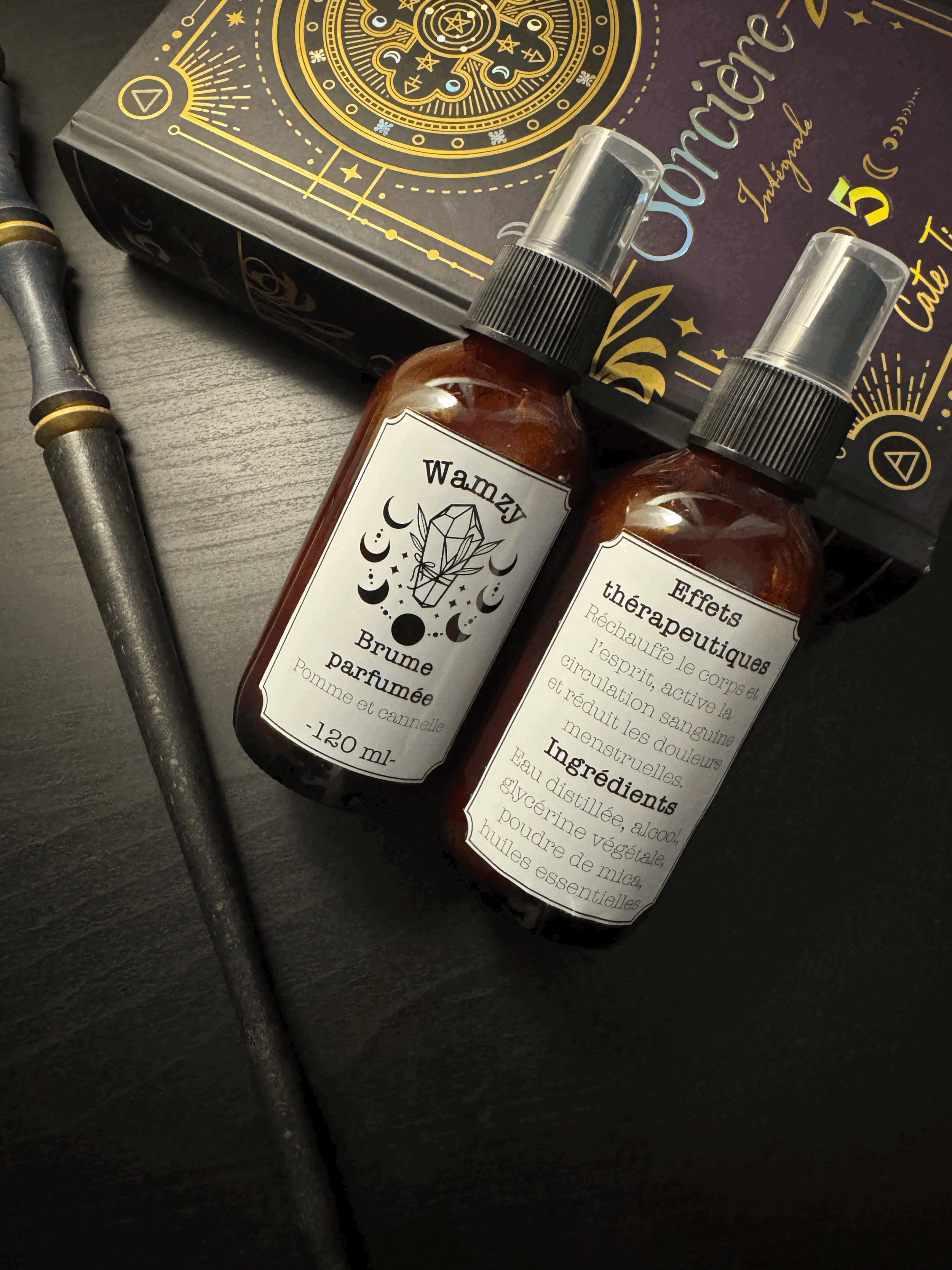 Deux flacons en verre avec étiquettes en français, un avec la mention 'Brume parfumée' à la pomme et cannelle, et l'autre avec 'Effets thérapeutiques' indiquant une utilisation pour la relaxation du corps et de l'esprit. À côté, un bâton de rançais et une boîte noire avec des dessins dorés et des textes en français.