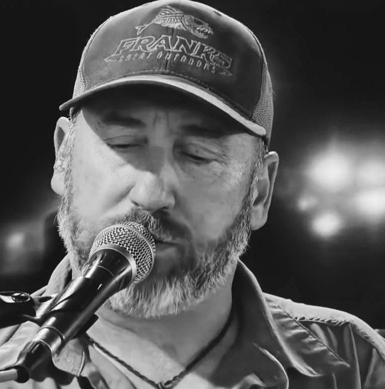 A bearded man with closed eyes singing into a microphone, wearing a cap with the logo 'Frank's Charter Outboards' in black and white.