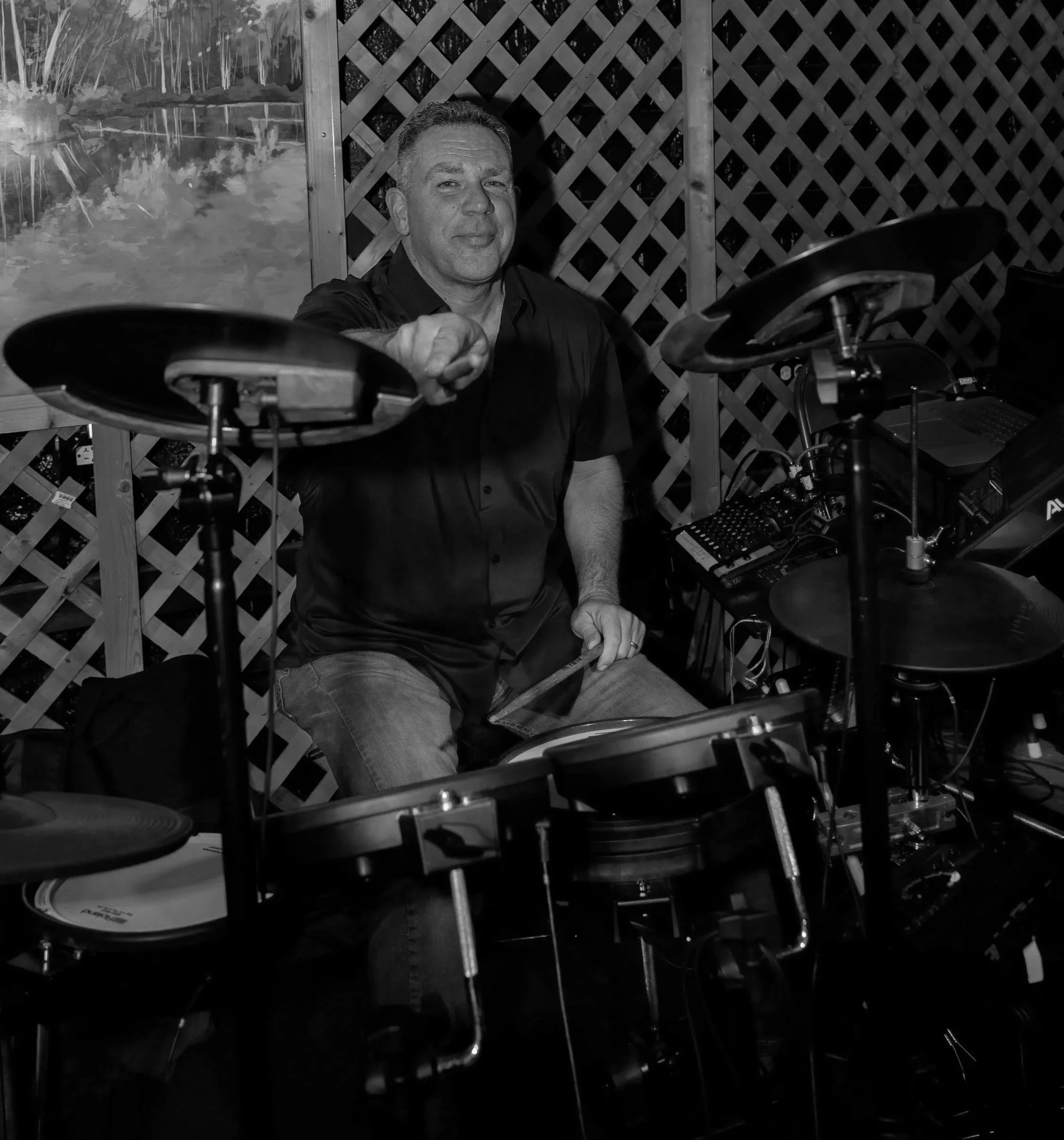 A man playing an electronic drum set in a room with lattice walls and a landscape painting on the wall.