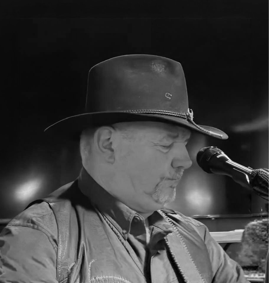 A man with facial hair wearing a wide-brimmed hat and a jacket, speaking into a microphone in a dimly lit setting.