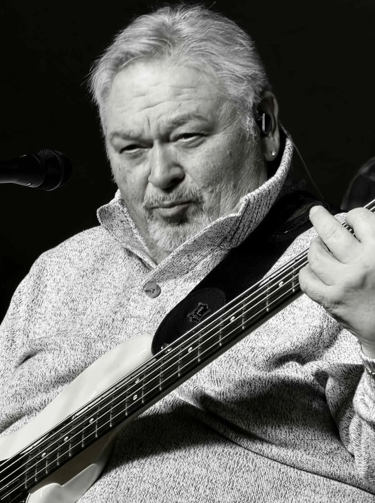 A black and white photo of a man with gray hair and a beard playing an electric guitar, wearing a zip-up sweater and an earpiece.