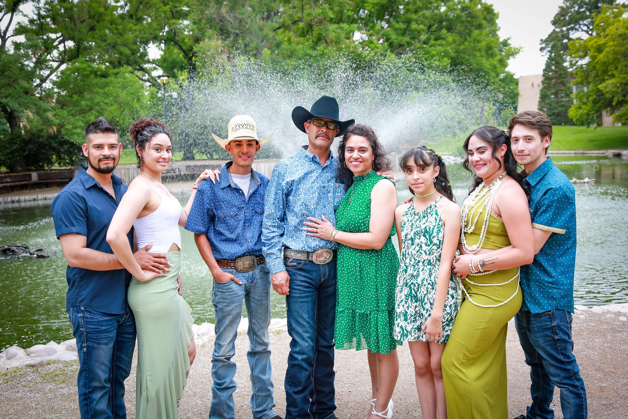 A diverse group of nine people standing in front of a pond with a fountain, surrounded by trees, smiling at the camera.