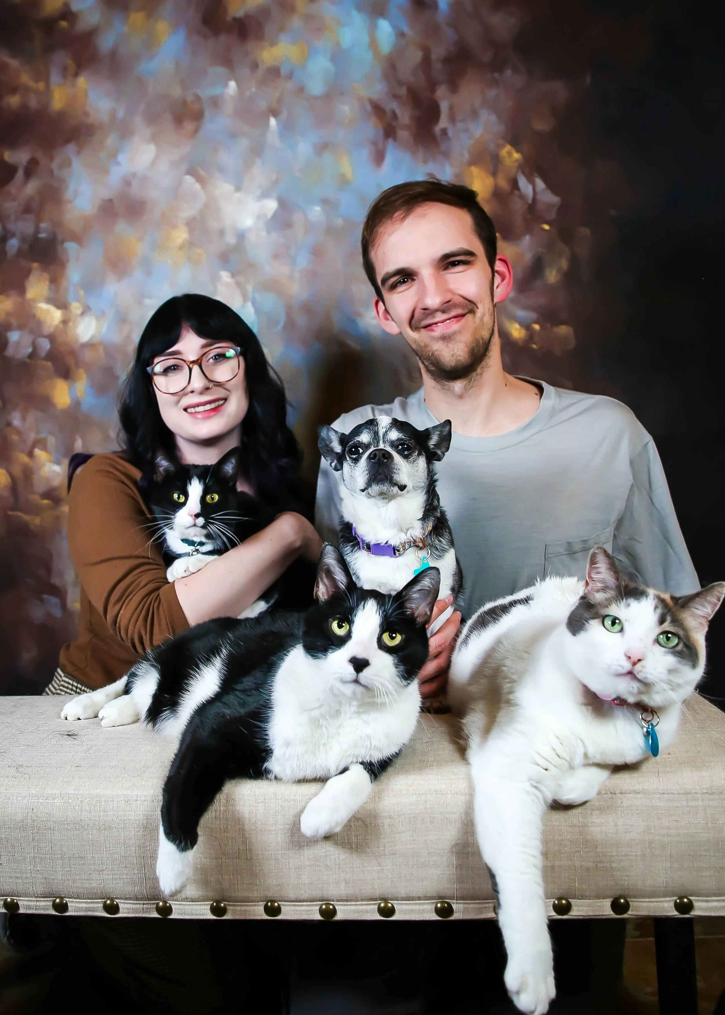 A smiling young man and woman with four pets: a black and white cat, a black and white dog, a small black dog, and a white and gray cat, in front of a painted background.