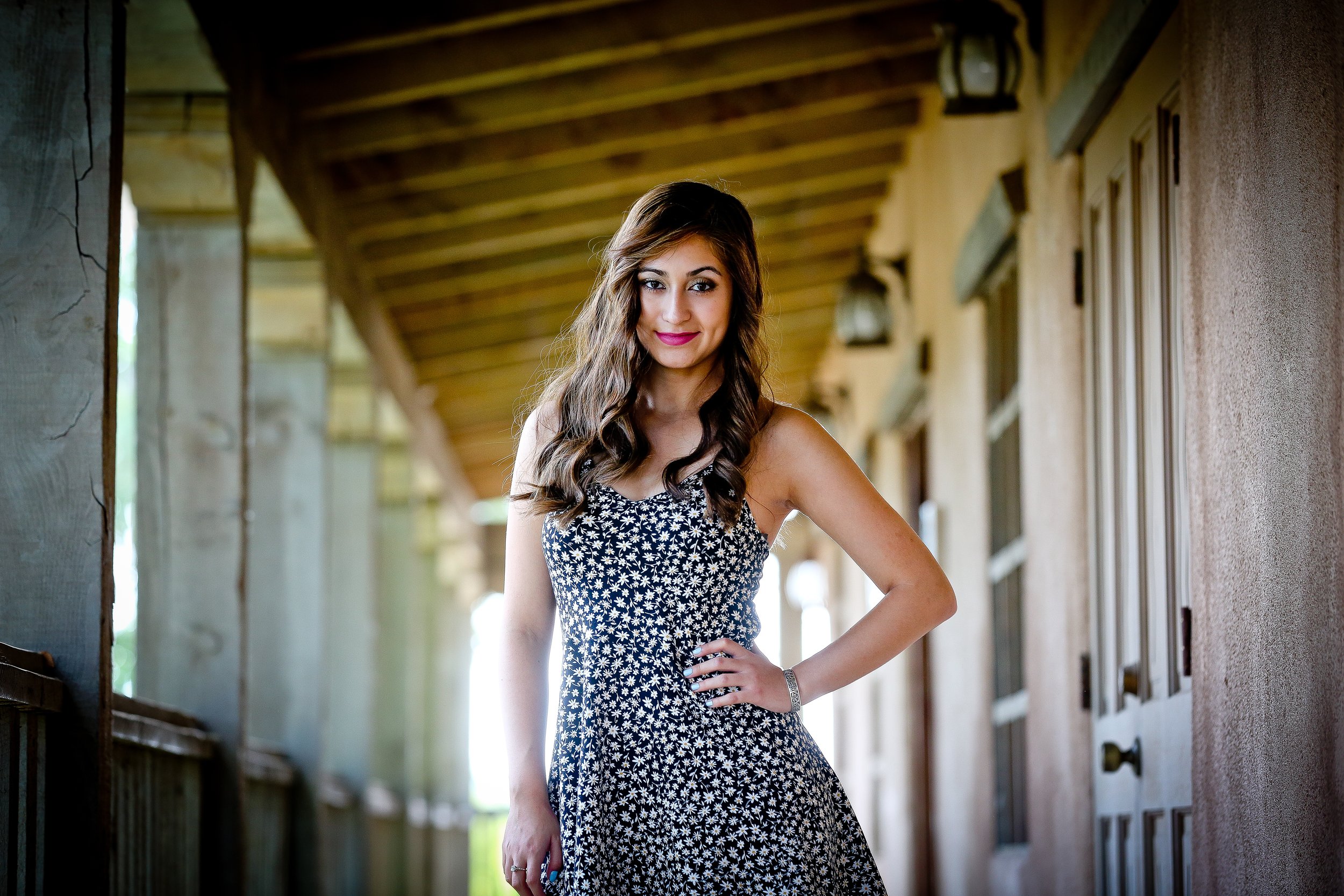 A young woman with wavy brown hair wearing a sleeveless, black and white floral dress, standing on a wooden porch with a playful expression, one hand on her hip, in a rustic setting with a wooden ceiling and walls.