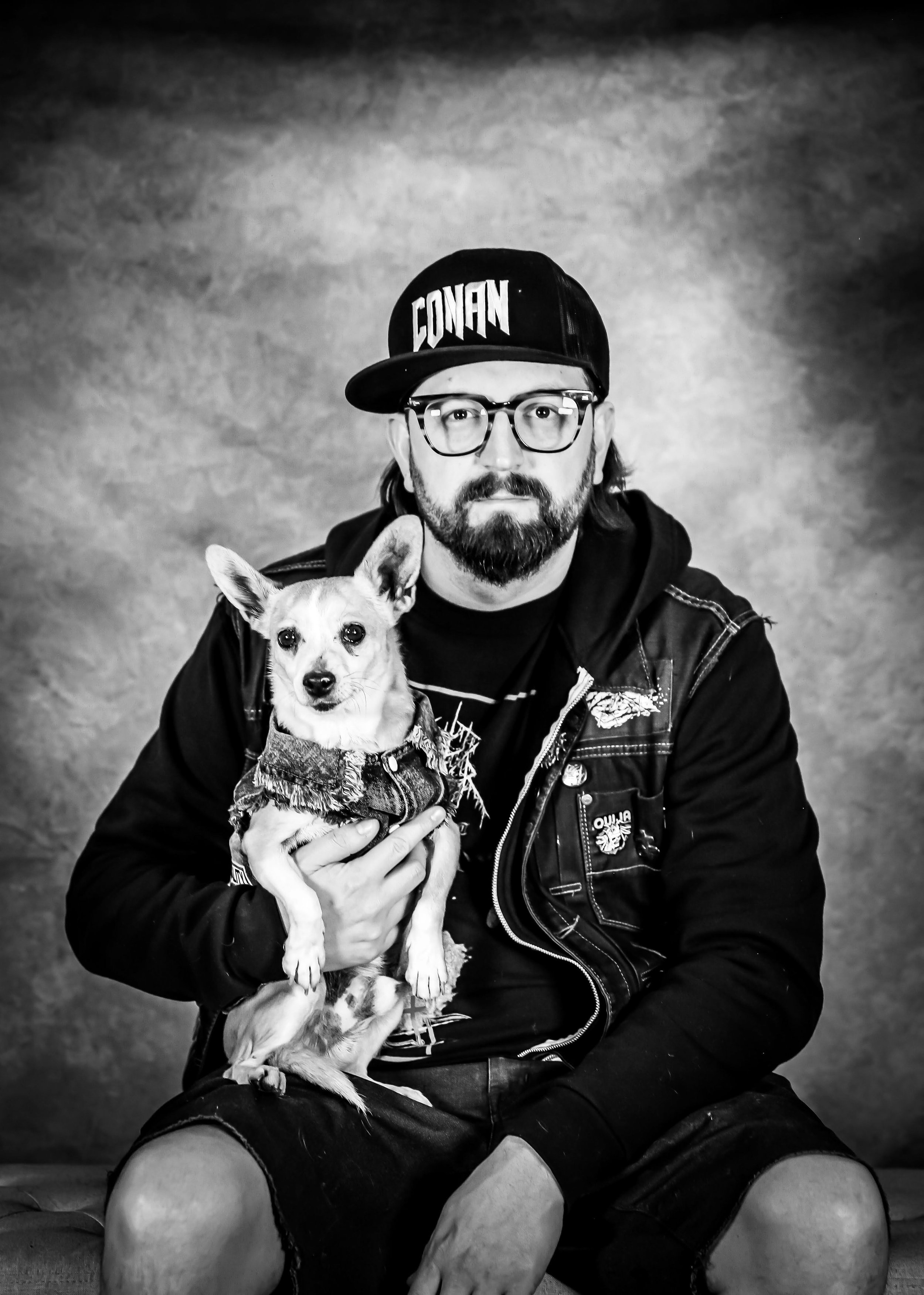 A man with glasses, a beard, and a cap that says "CONAN" sits holding a small dog wearing a denim vest, against a textured background.
