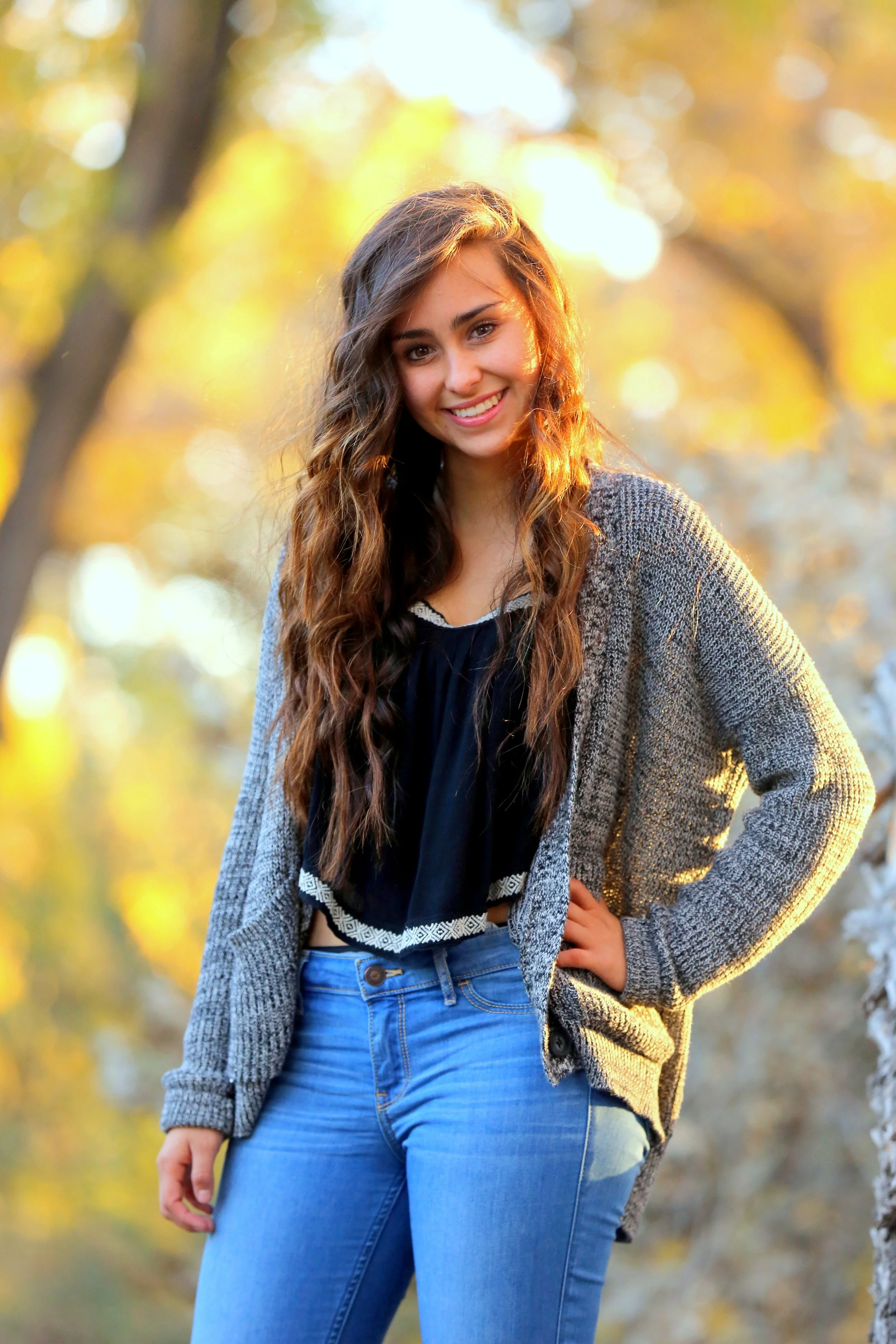 A young woman with long, wavy brown hair smiles at the camera. She is wearing a gray knitted cardigan, a dark blue top with white trim, and light blue jeans. The background features trees with yellow and orange autumn leaves, illuminated by warm sunlight.