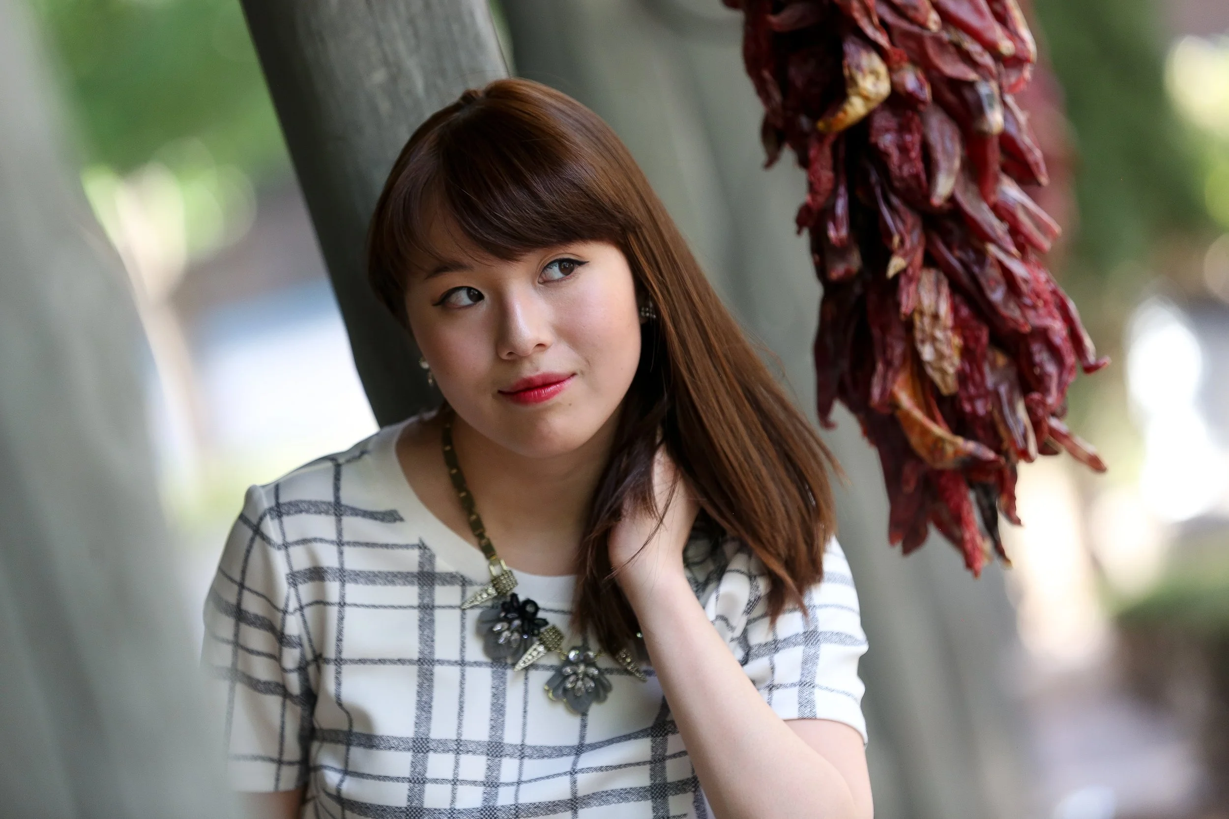 A young woman with brown hair, wearing a white checkered shirt and a floral necklace, standing near hanging red chili peppers.