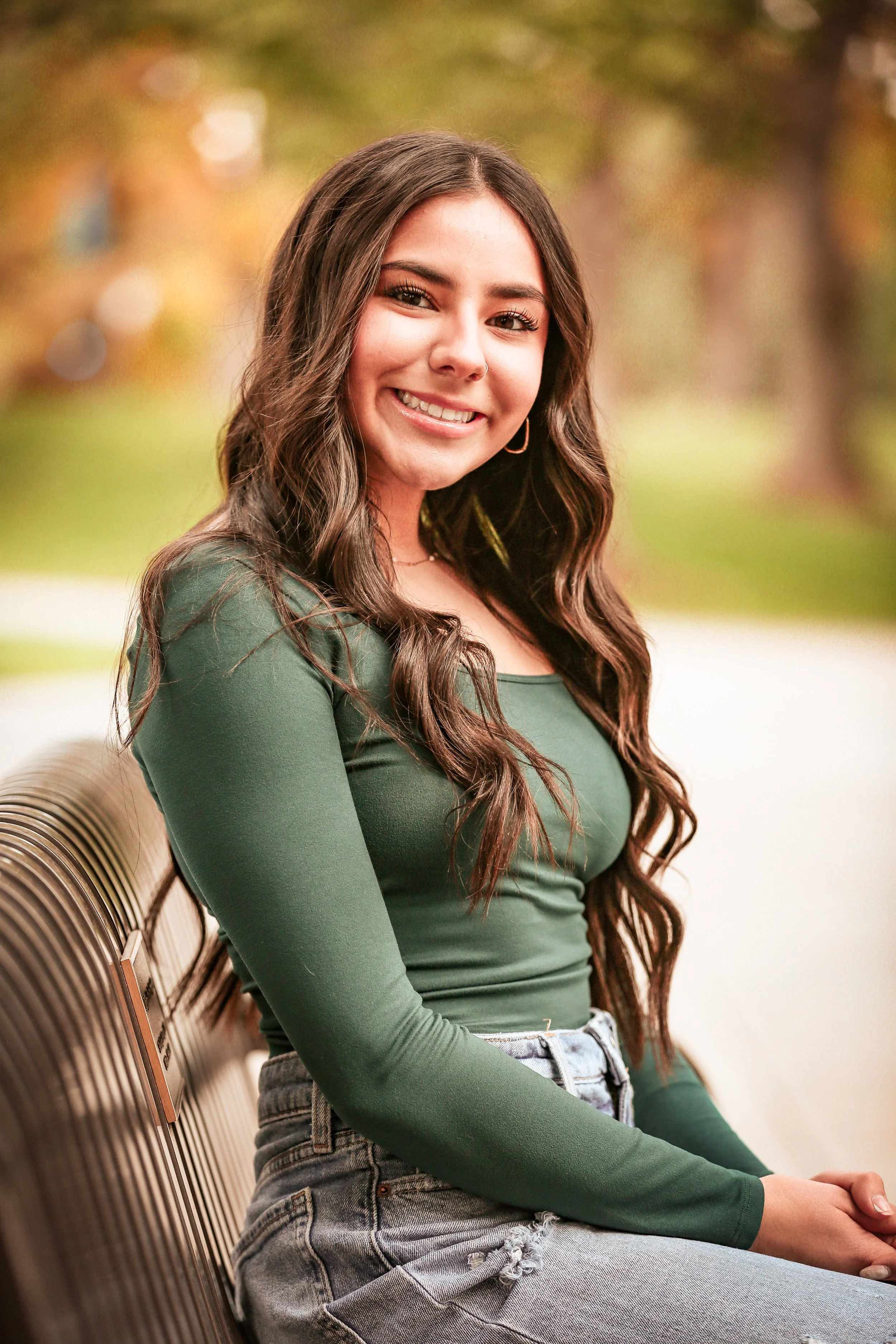 A young woman with long wavy brown hair sitting on a park bench, smiling at the camera, wearing a green long-sleeve shirt and ripped jeans, with trees blurred in the background.
