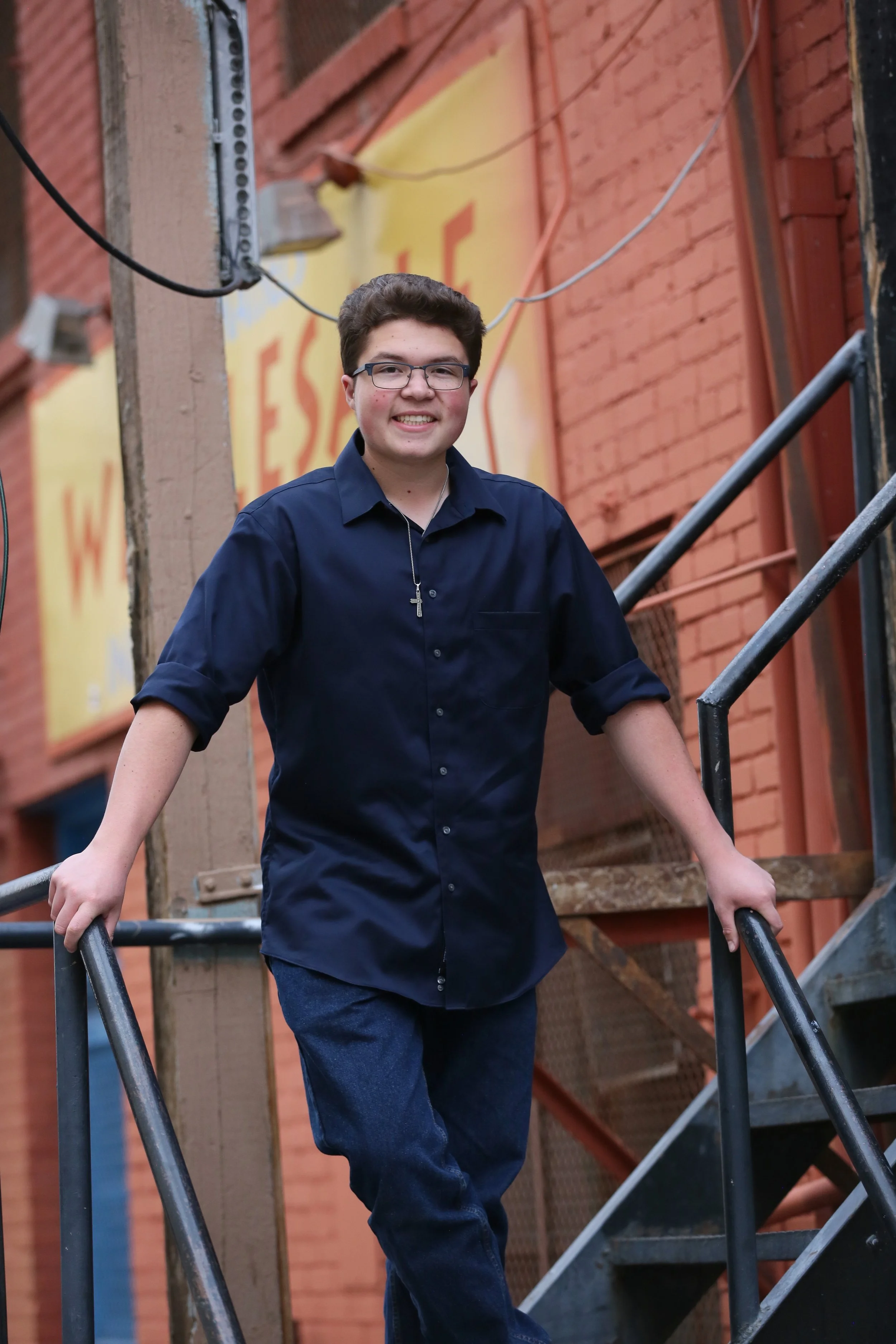 A young man with glasses and a cross necklace, wearing a navy blue button-up shirt and jeans, smiling while standing on a metal staircase outside a brick building.