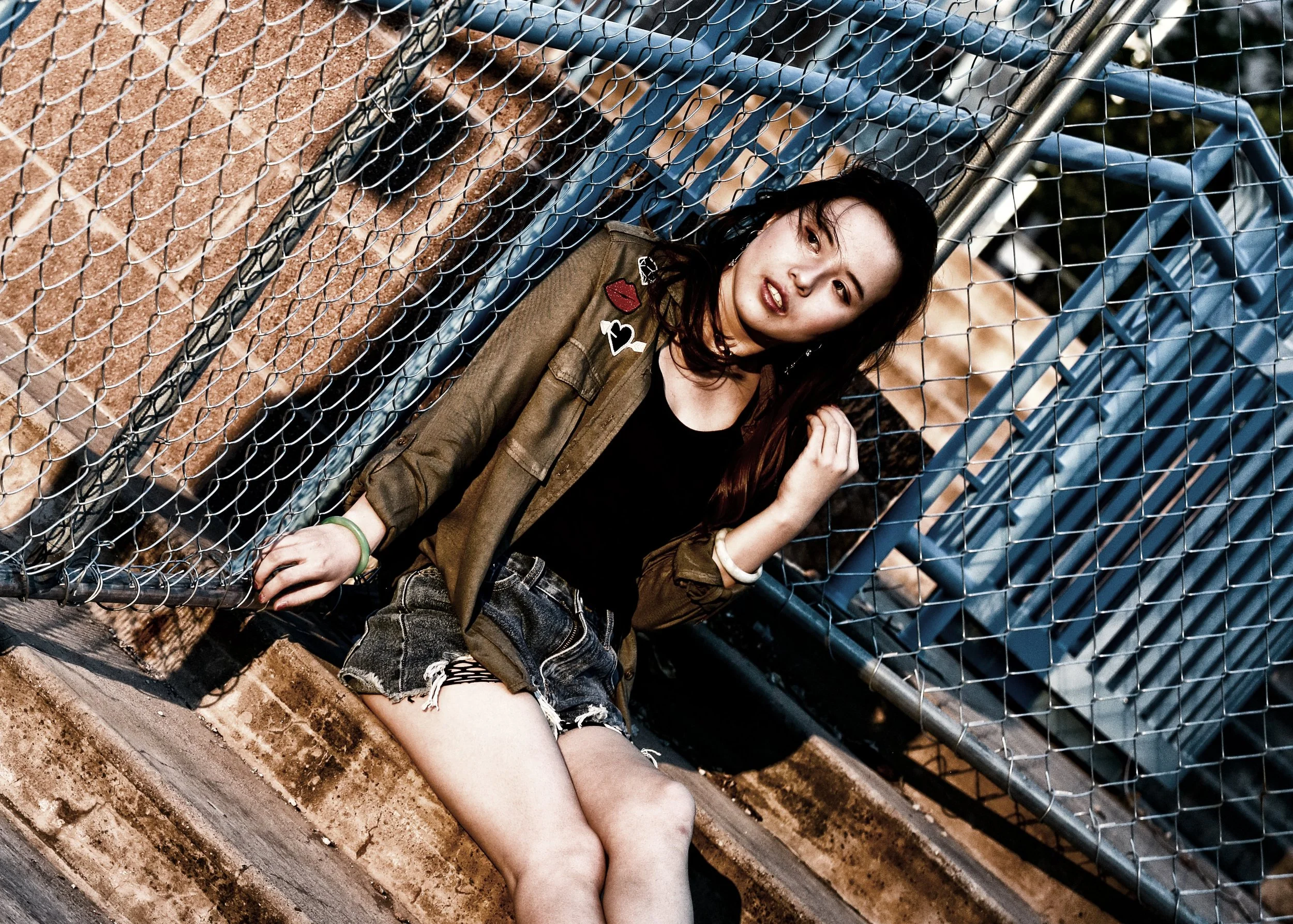 A young woman with dark hair and a casual style is sitting by a chain-link fence, with some graffiti behind her and blue metal bleachers in the background. She is wearing a brown jacket with patches, a black top, and ripped denim shorts.