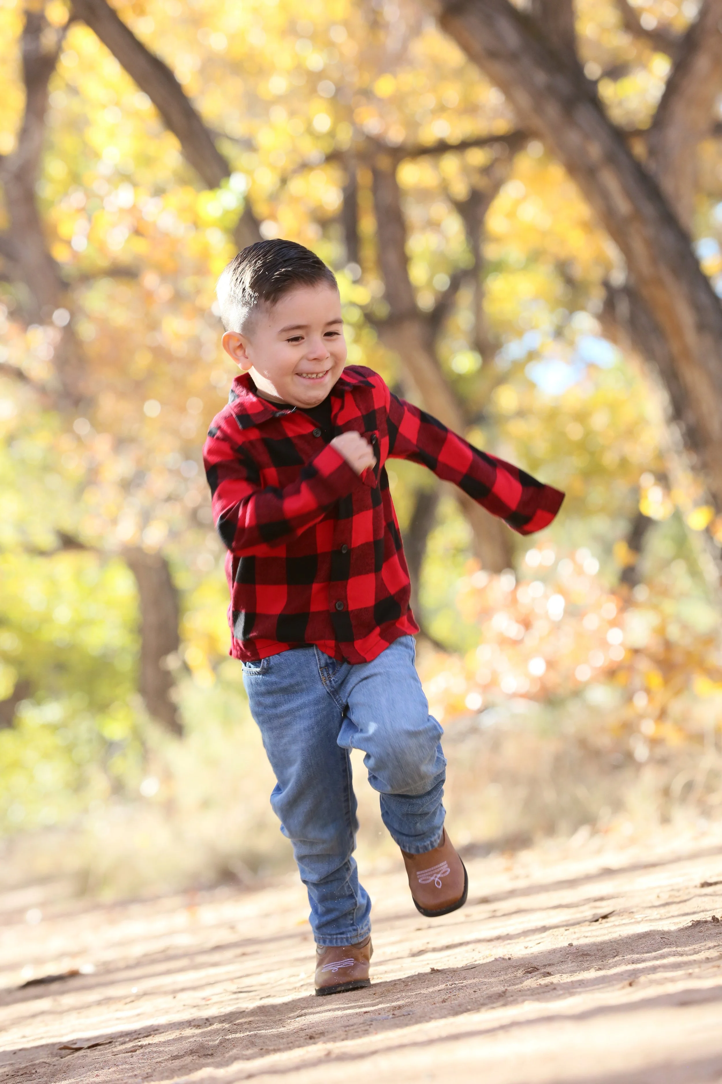 A young boy wearing a red and black checkered shirt, blue jeans, and brown shoes running outdoors on a dirt path surrounded by autumn foliage.