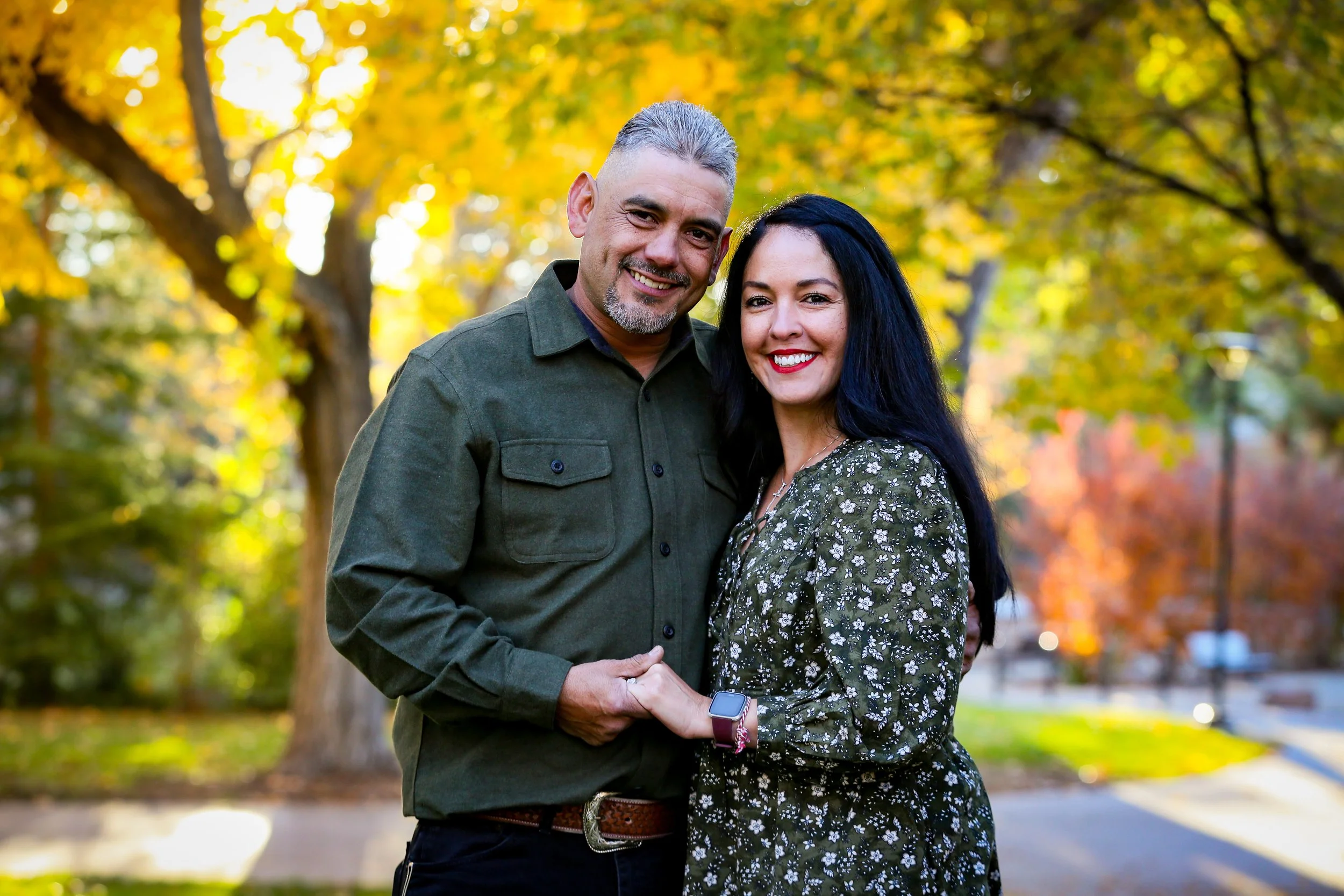 A smiling middle-aged man and woman stand close together outdoors in a park during fall, with yellow and orange leaves on trees in the background, holding hands and looking at the camera.
