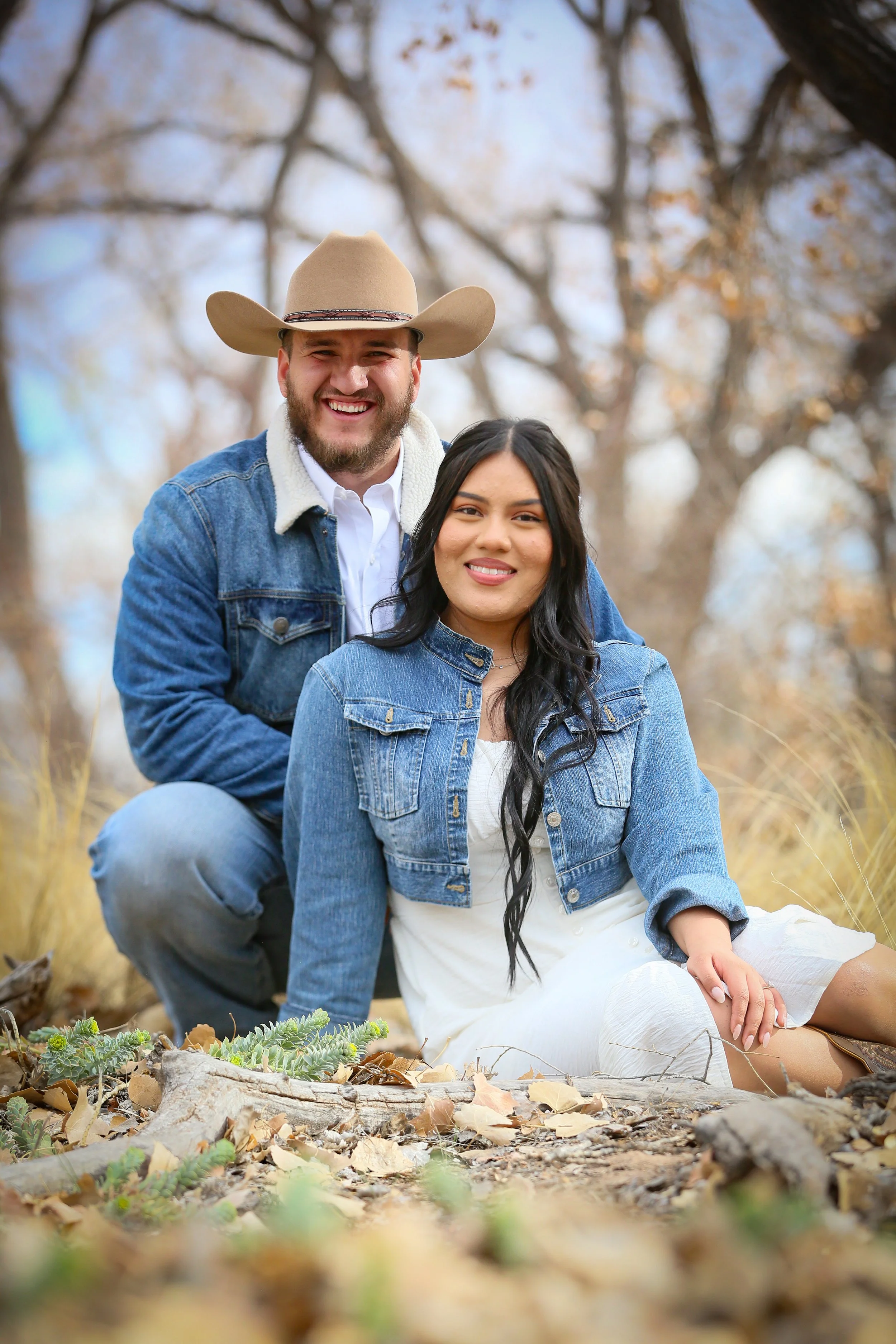 A smiling couple outdoors in a fall setting, sitting on the ground with fallen leaves, surrounded by trees with autumn foliage, wearing denim jackets.