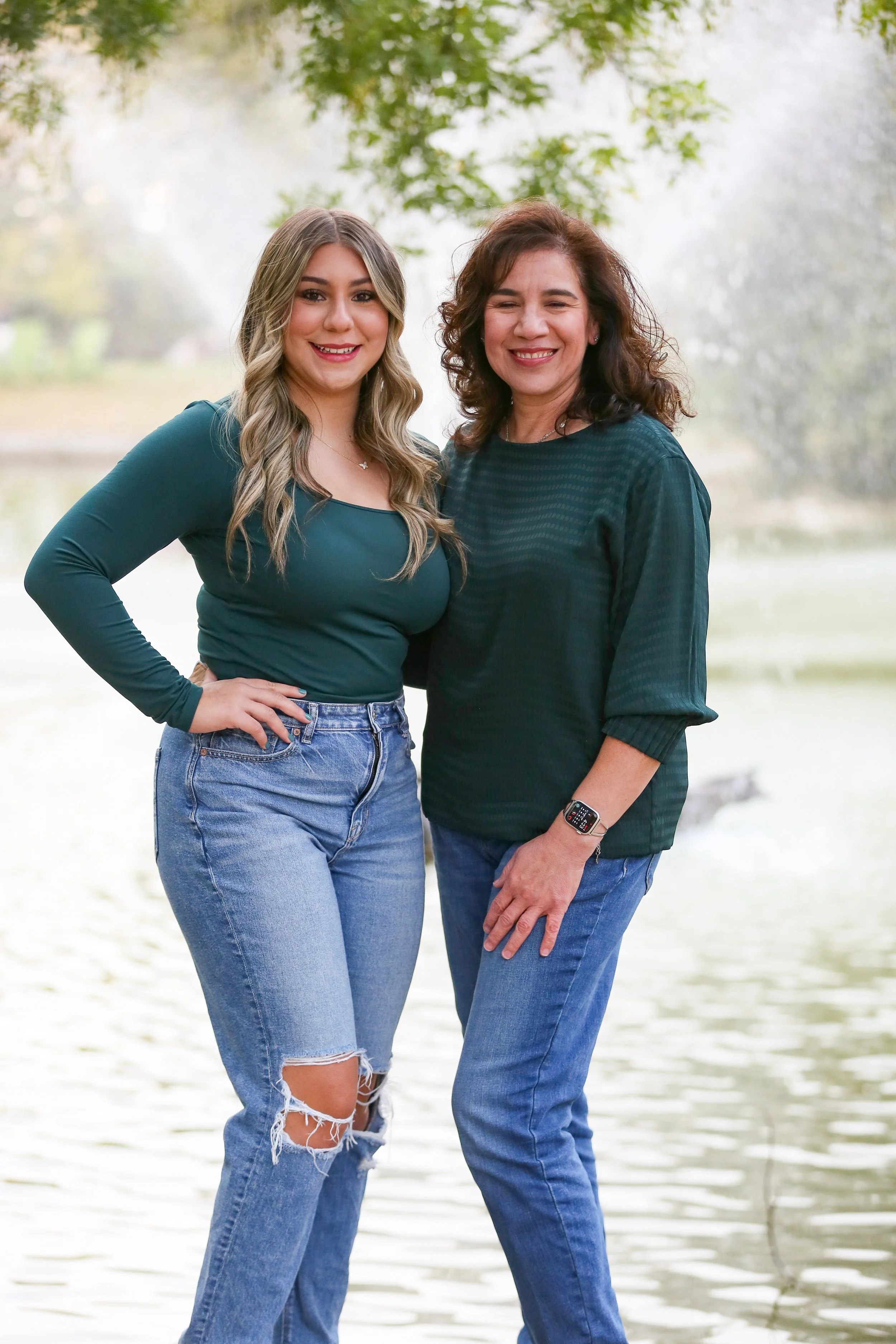 A young woman and an older woman standing close together outdoors near a body of water and trees, smiling at the camera.
