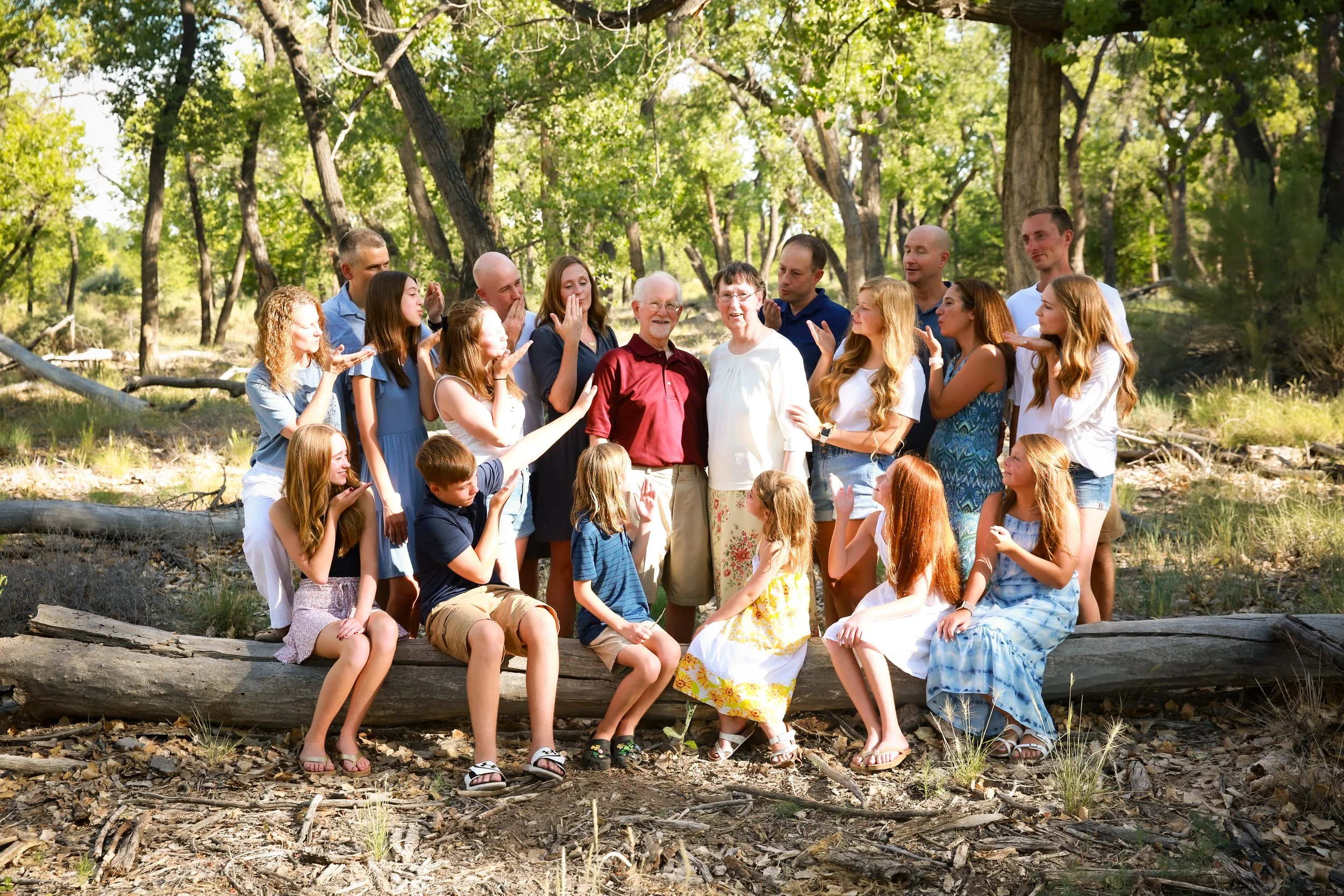 A large group of people, including adults and children, gathered outdoors in a wooded area, smiling and posing for a photo.