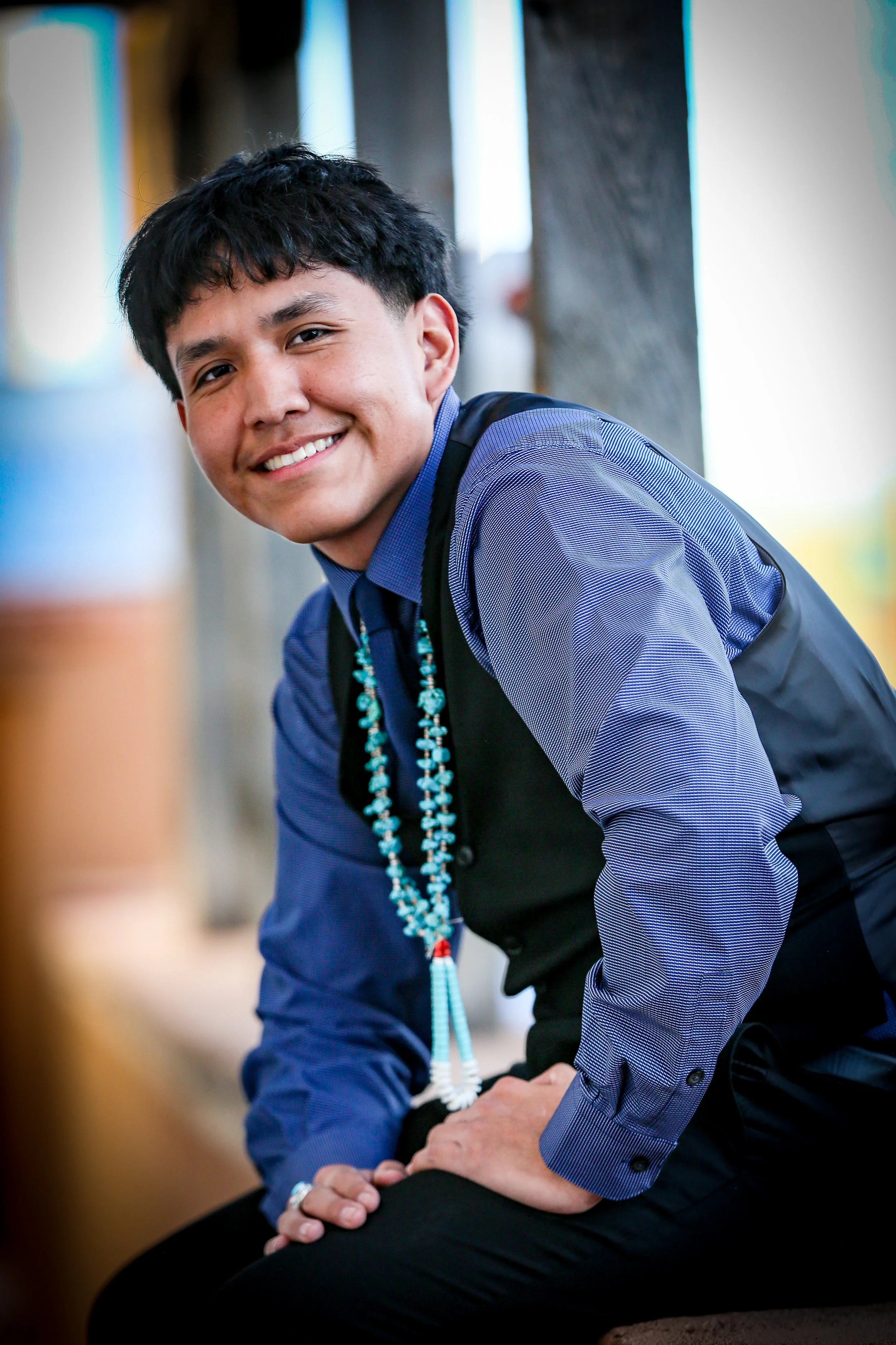 A young man with dark hair, wearing a blue shirt and black vest, is sitting and smiling at the camera. He has turquoise and white beaded necklaces around his neck and appears to be indoors with blurred background.