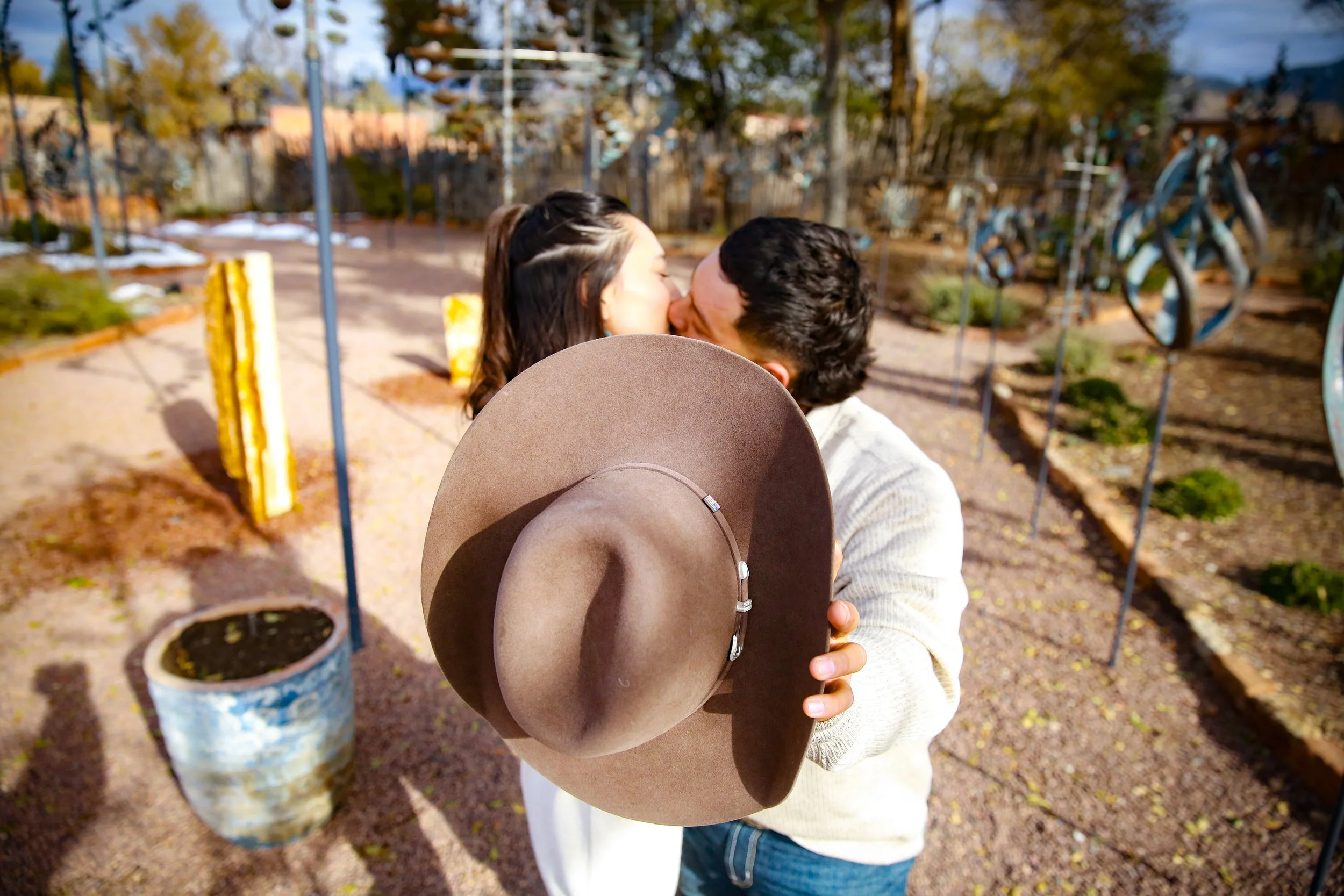 A couple kiss outdoors, with the man holding a wide-brimmed brown hat. The background features a garden with colorful art sculptures and potted plants.
