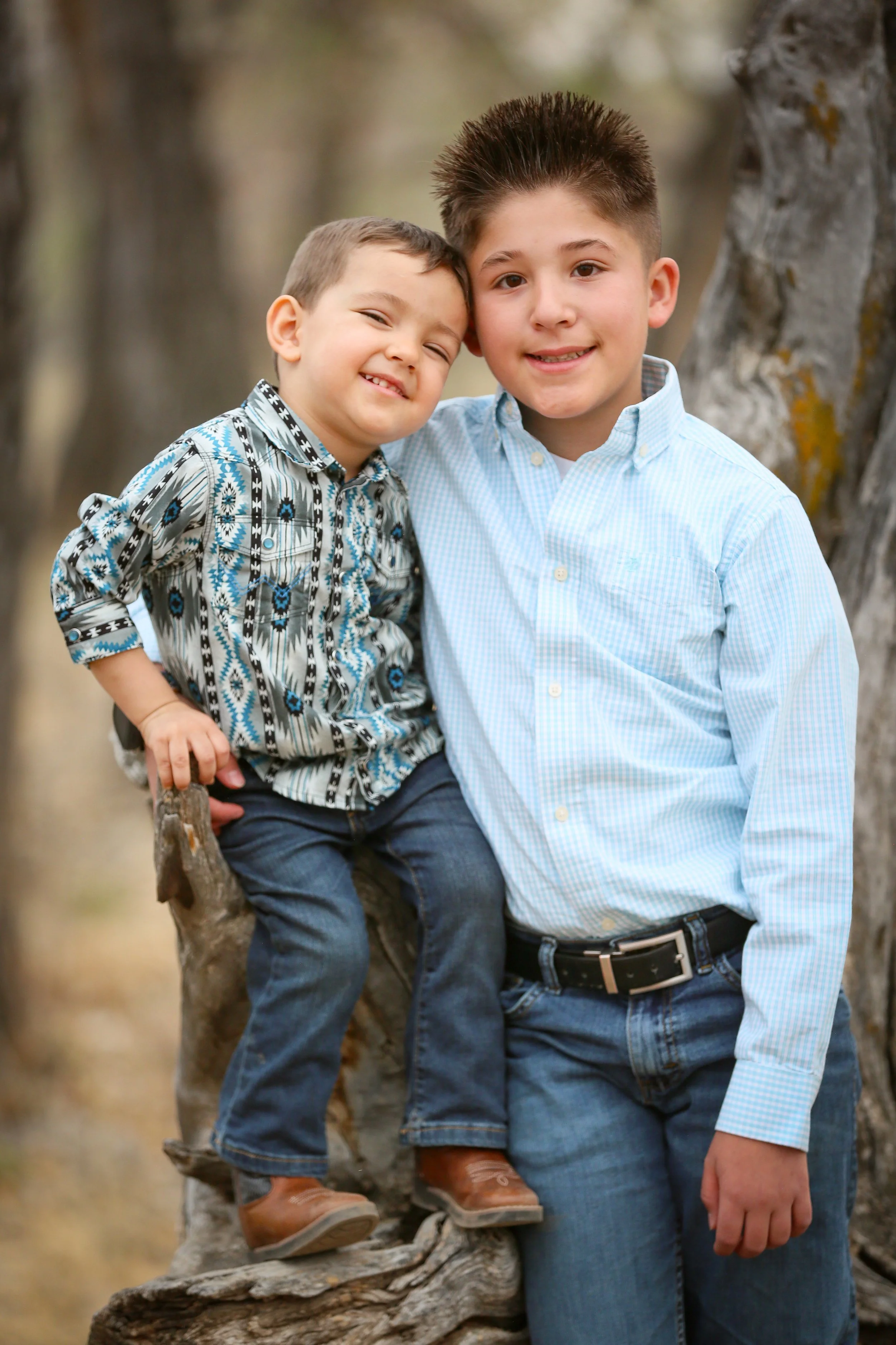 Two boys, one older and one younger, standing outdoors near a tree. The older boy has short brown hair and is wearing a light blue button-up shirt and jeans. The younger boy has short light brown hair and is wearing a patterned long-sleeve shirt, jeans, and brown shoes. The younger boy is sitting on a tree branch while the older boy stands beside him, both smiling at the camera.