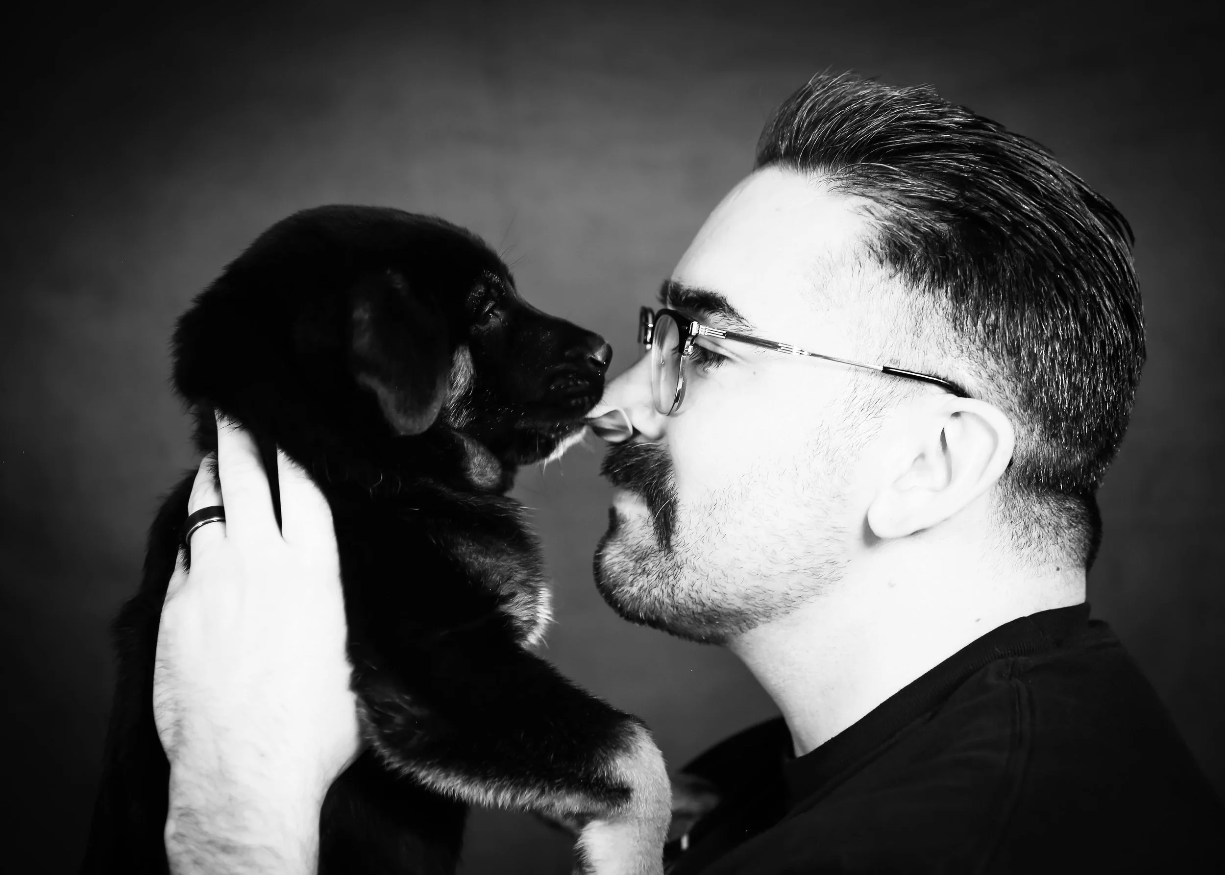 A man wearing glasses holding a black and tan puppy close to his face touching noses in a black and white photo.