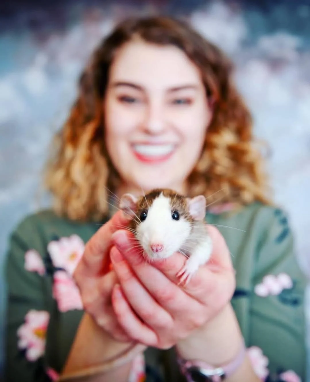 A woman with wavy brown hair smiling and holding a small, white and brown hamster in her hands, with a blurred background.