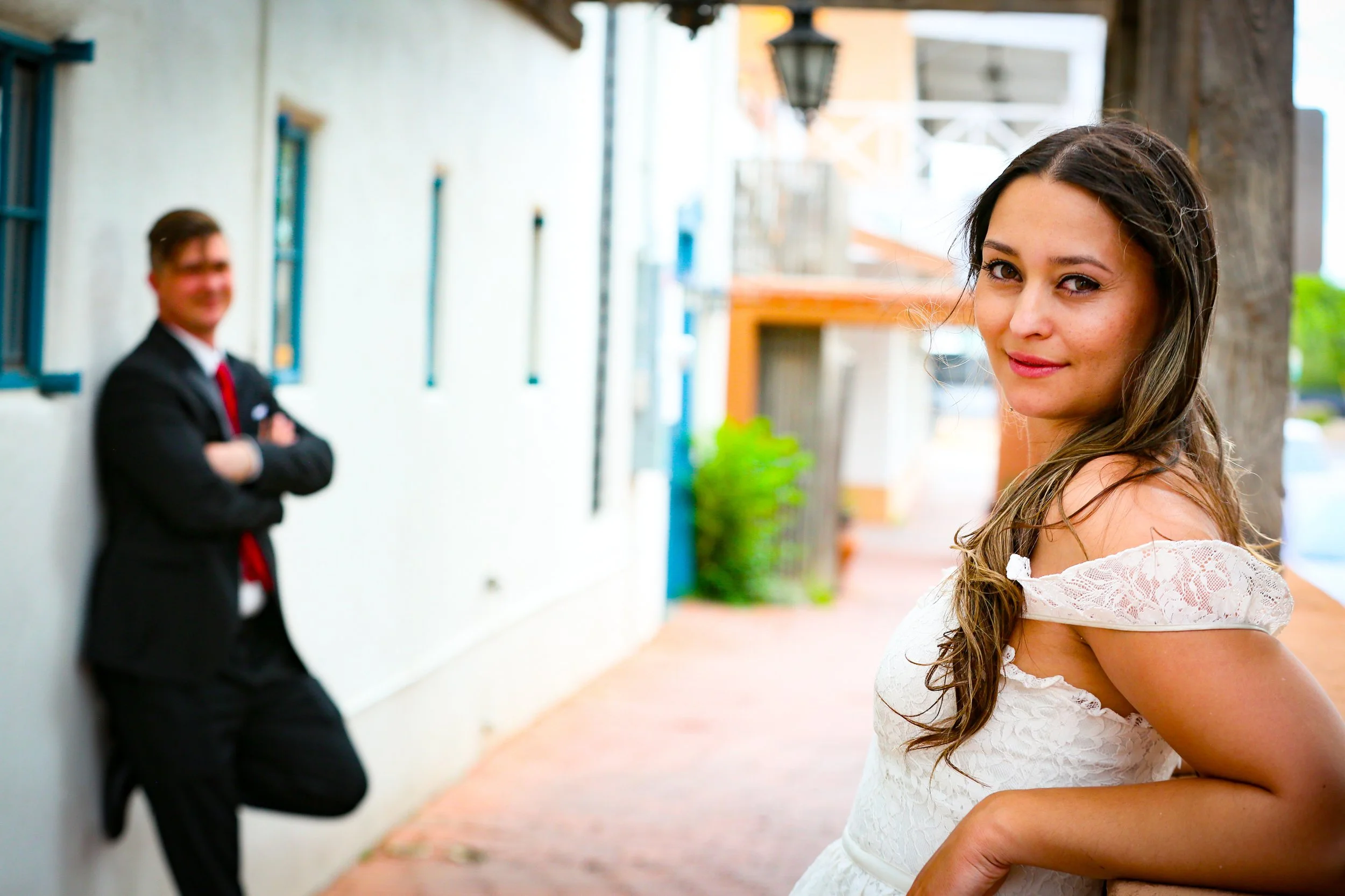 A woman in a white lace dress smiling at the camera, standing on a sidewalk, with a man in a suit leaning against a wall in the background.